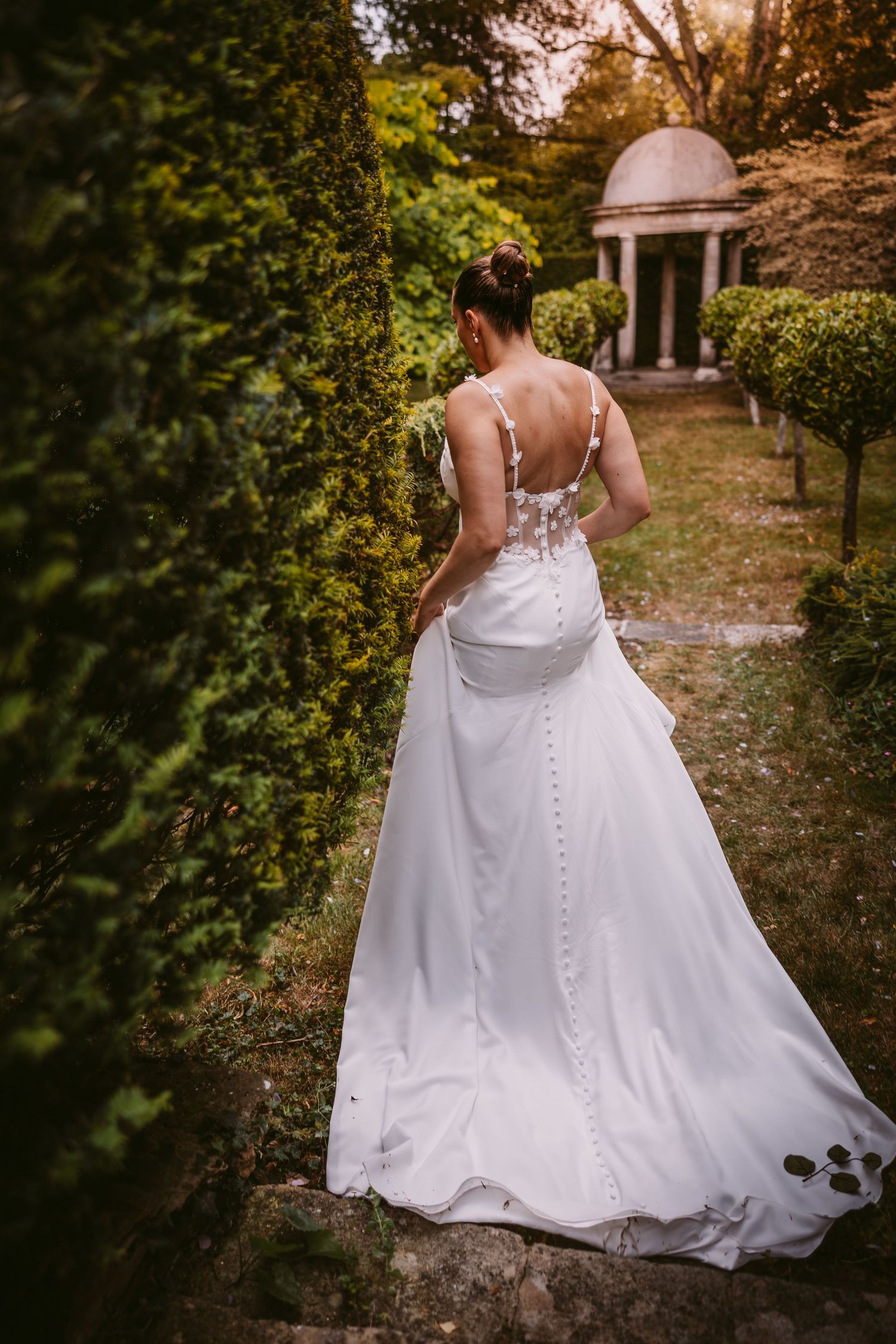 A bride in a white wedding gown with button details walks through a garden path during golden hour.