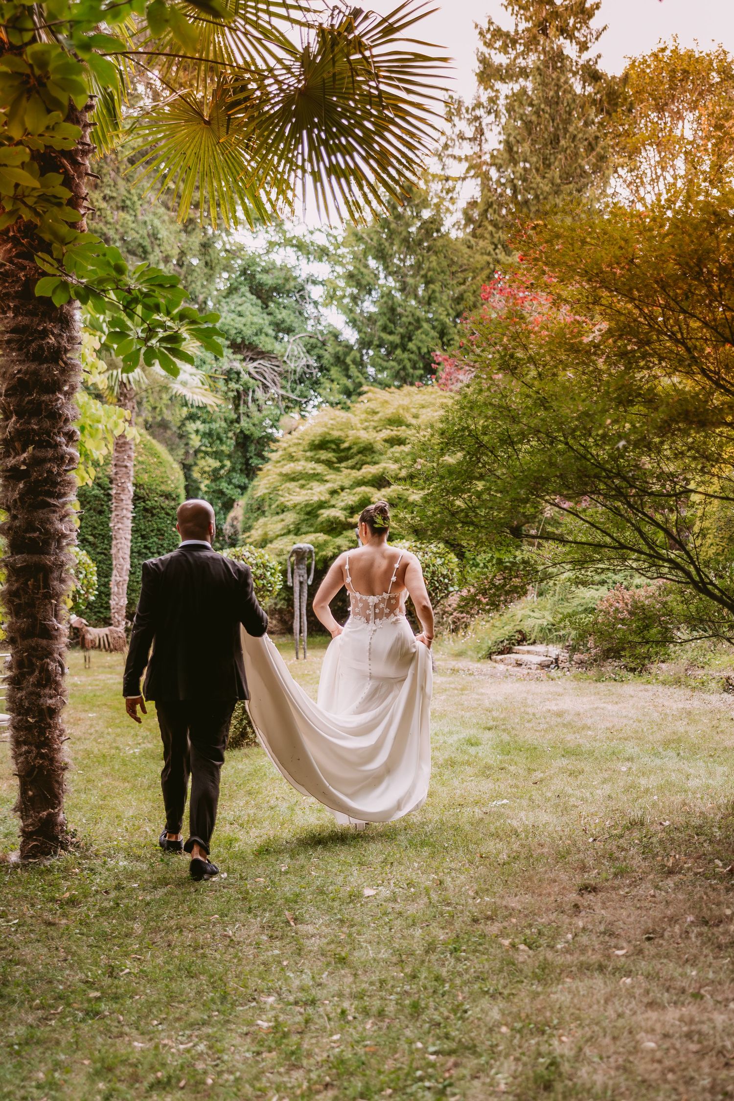 A couple in formal wedding attire walks through a scenic garden path surrounded by autumn foliage and palm trees.