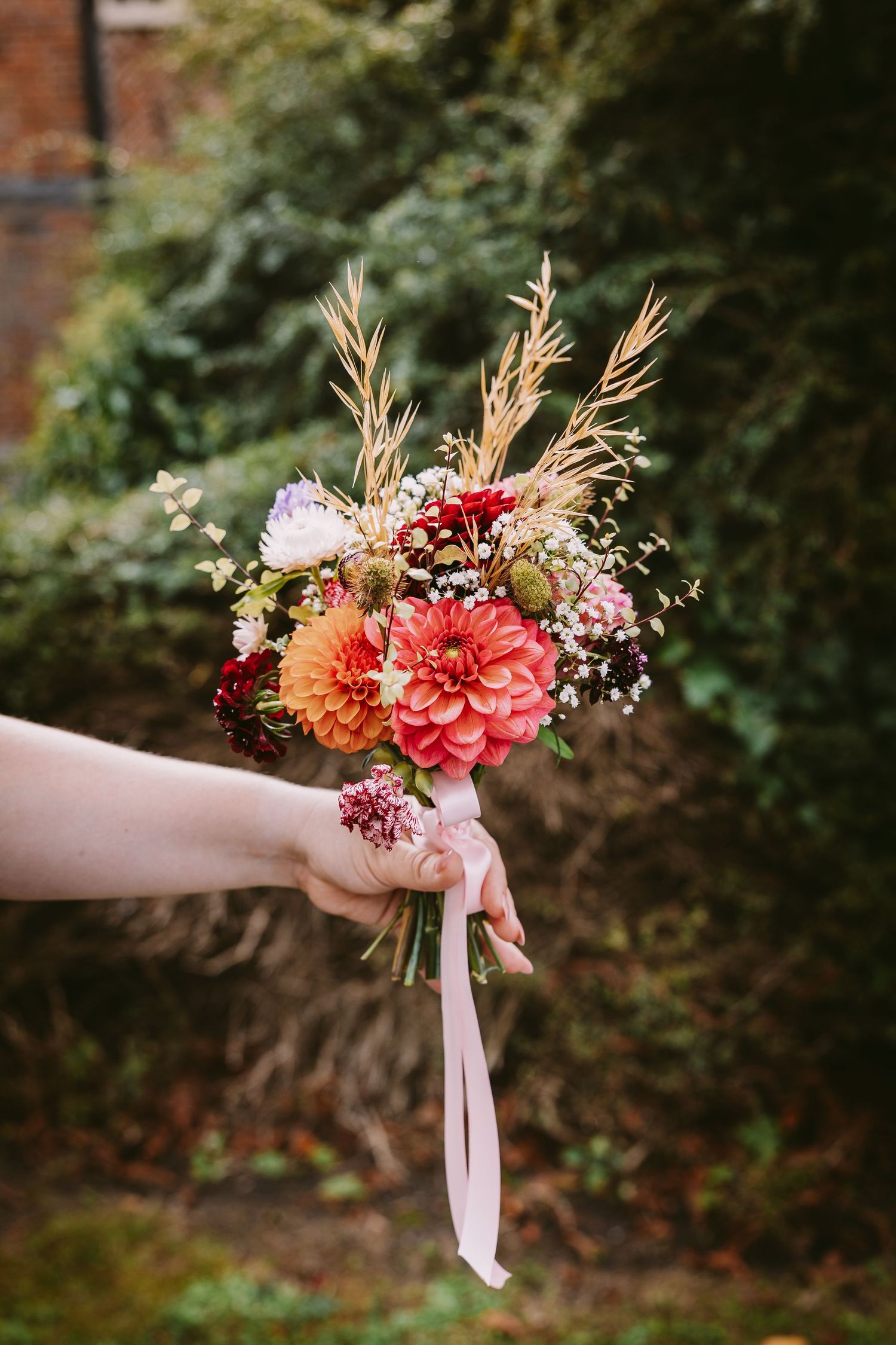 A hand holds a romantic bouquet of pink and coral dahlias with dried grasses tied with a light pink ribbon.