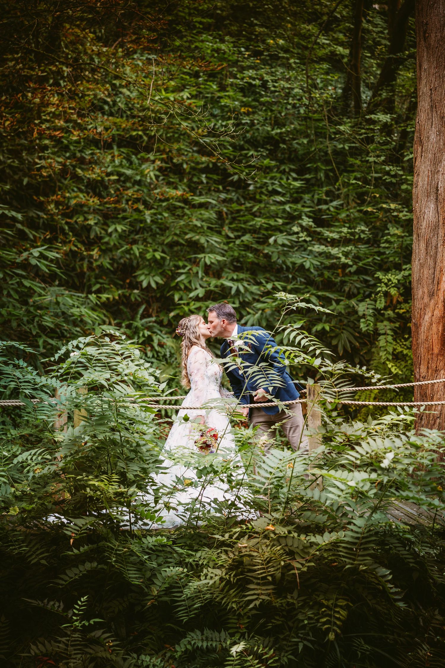 A romantic wedding couple shares an intimate moment surrounded by lush green ferns in a forest setting.