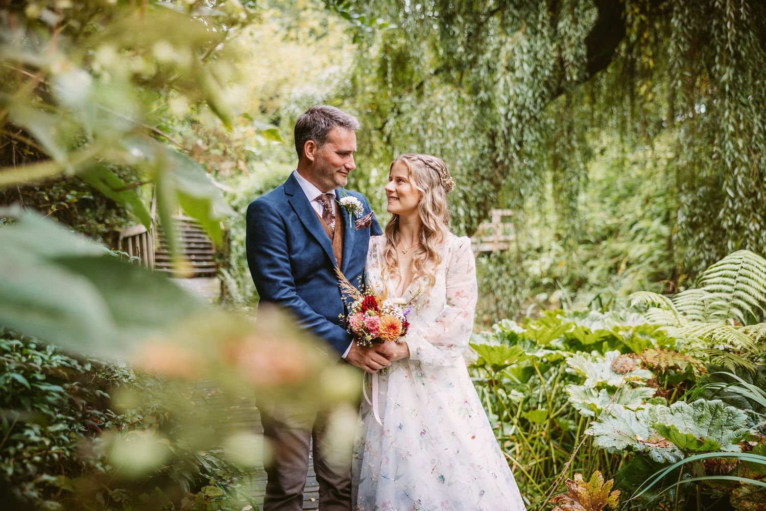 A romantic couple in wedding attire embraces in a lush garden setting with soft natural lighting and greenery.