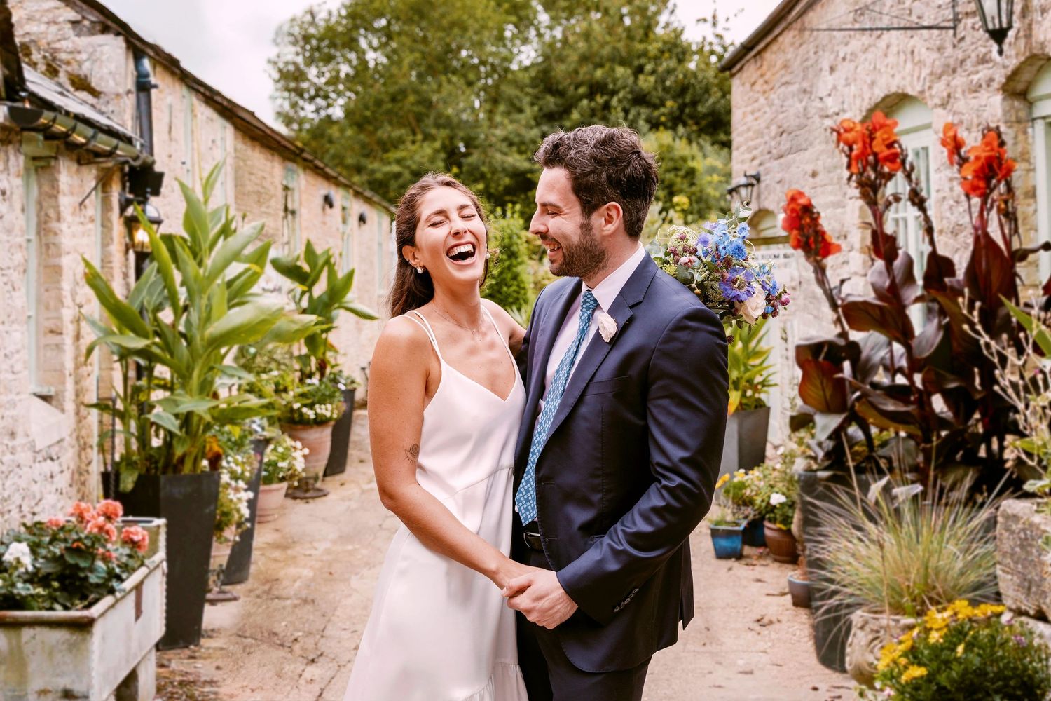 A joyful newlywed couple share a candid moment together in a garden walkway surrounded by lush plants.
