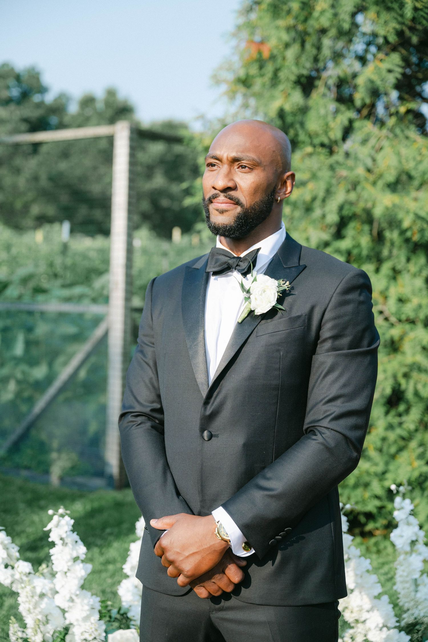 Groomsmen in black tuxedos line up outdoors with white floral arrangements at an elegant garden wedding ceremony. - Elawa Farm