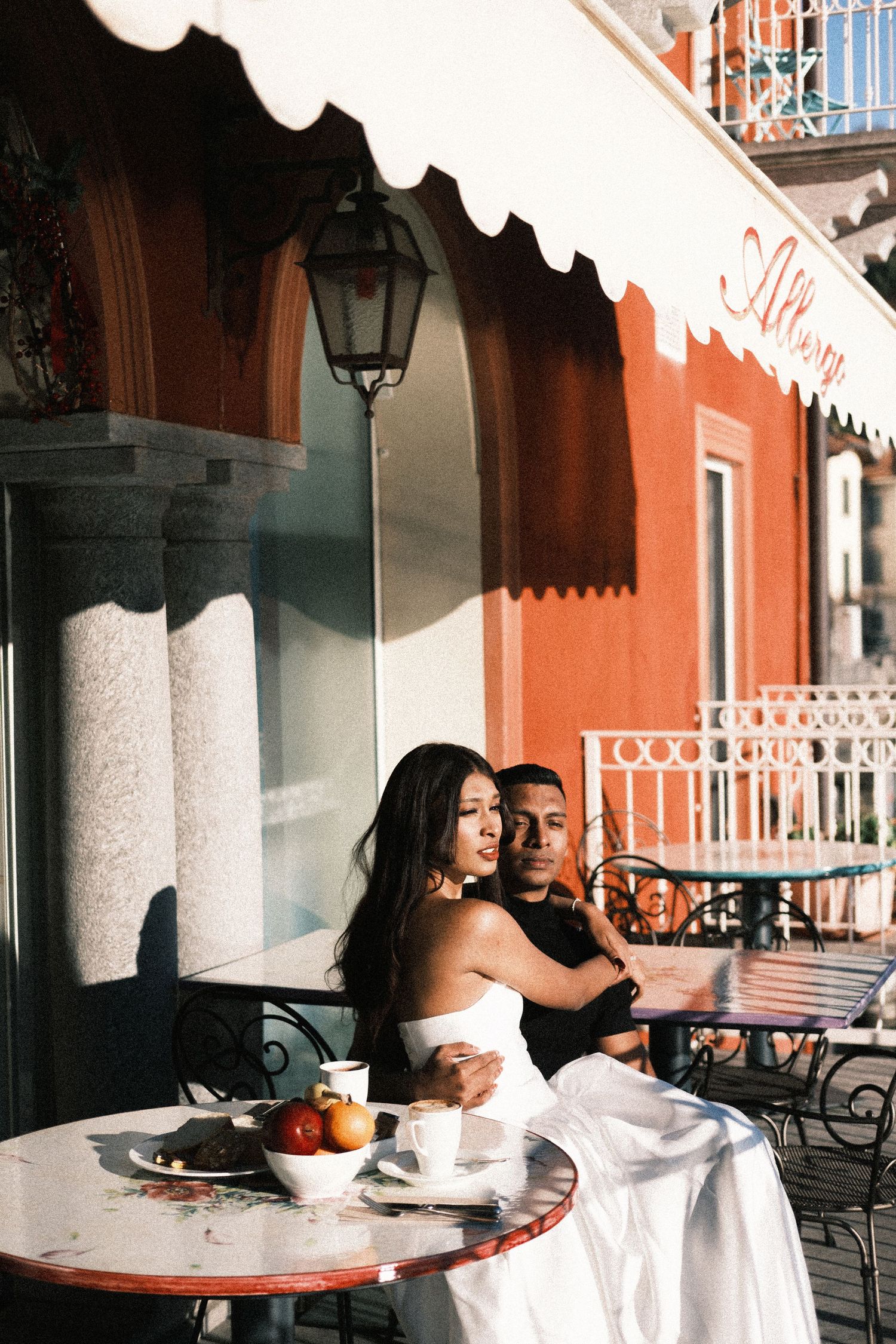 Couple enjoying coffee at an outdoor cafe table with red building facade and ornate lamp post in background.