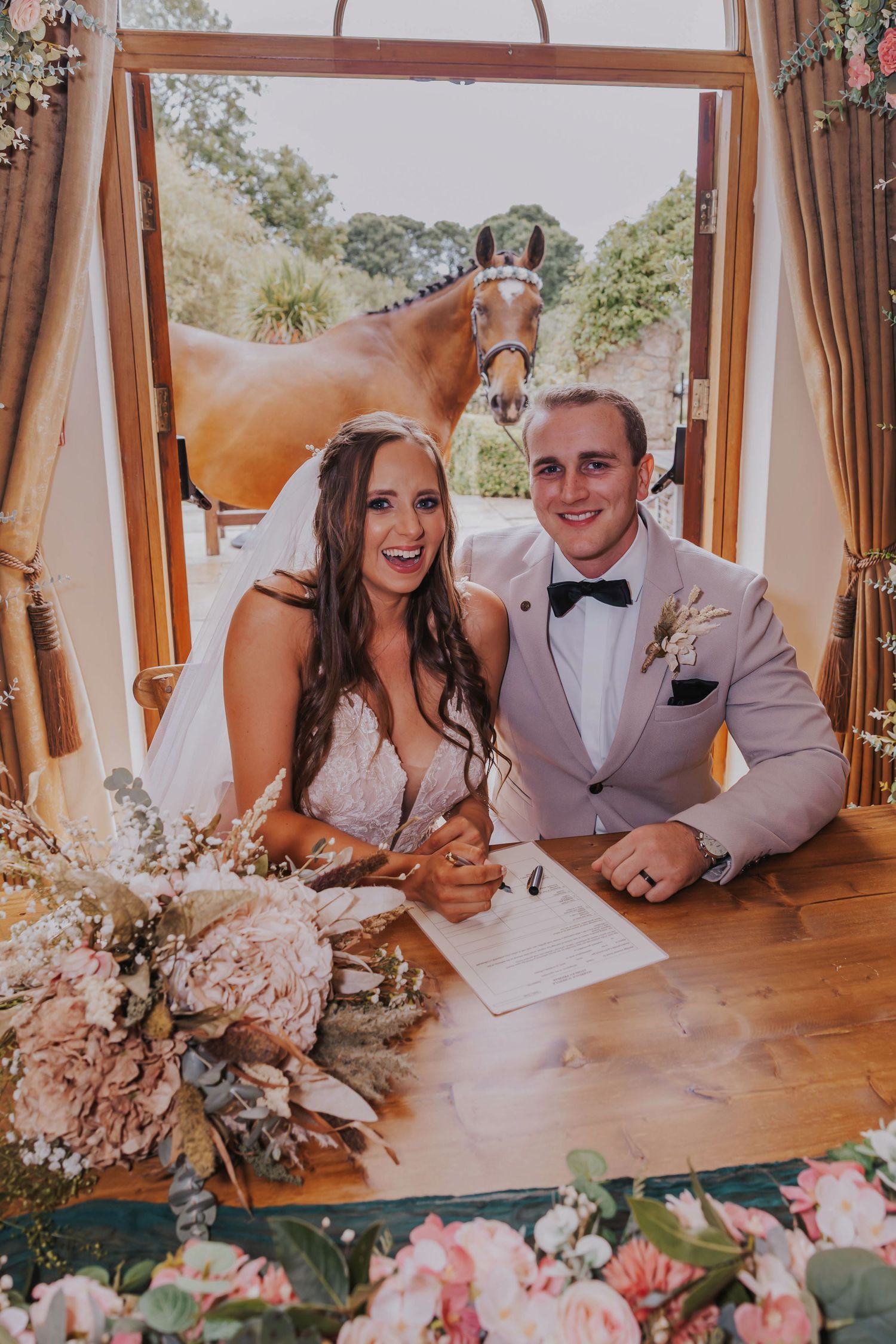 Happy couple signing marriage documents at a rustic wooden table decorated with pink floral arrangements.