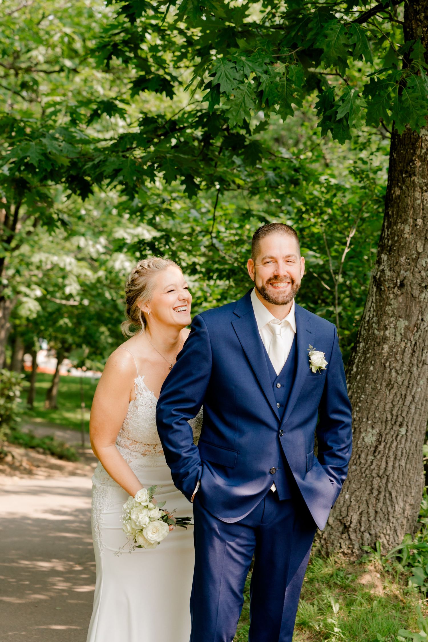 Smiling bride and groom in navy suit walking together on a tree-lined path on their wedding day.