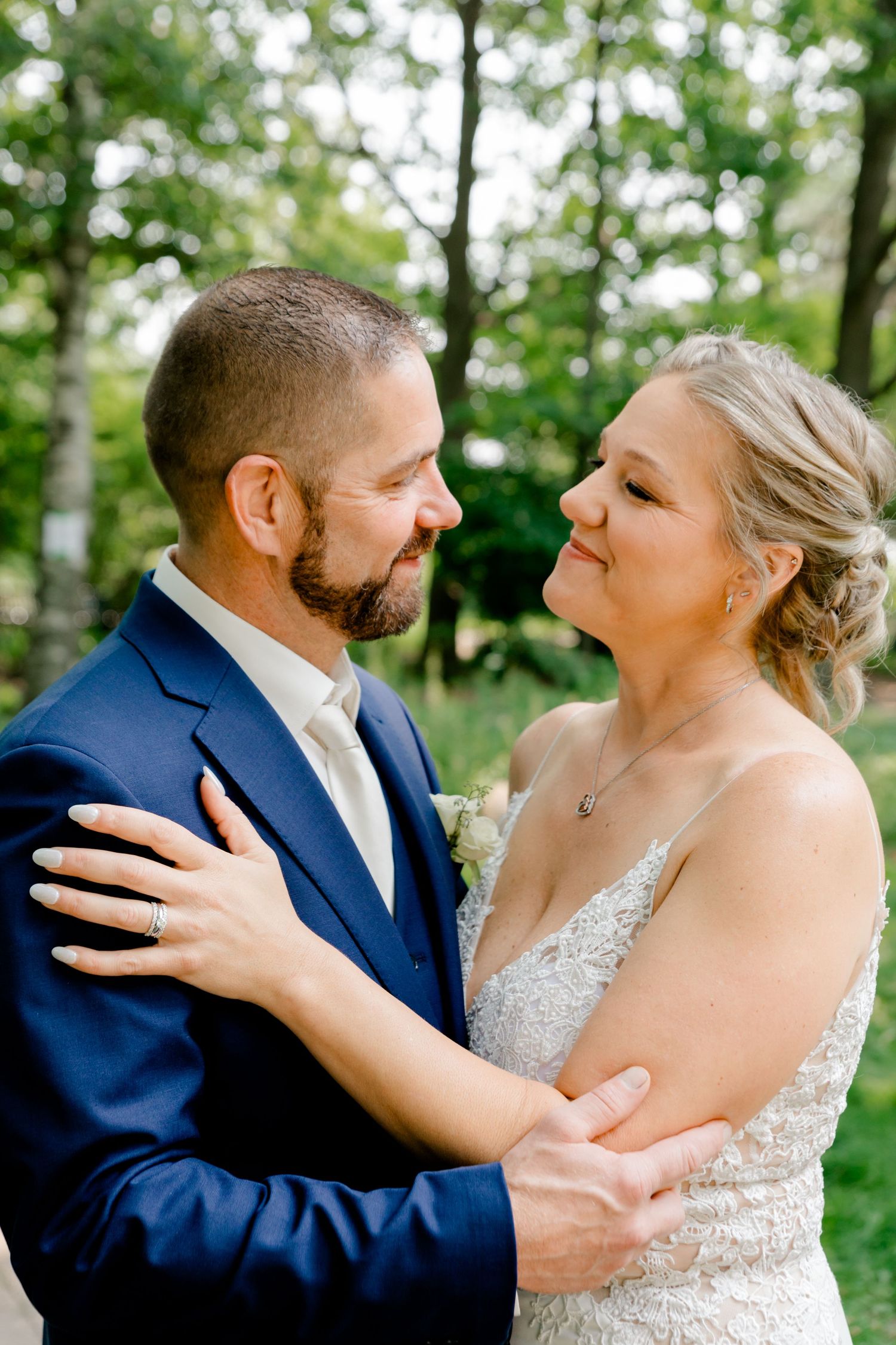 Bride and groom gazing lovingly at each other while embracing outdoors in a lush green setting.