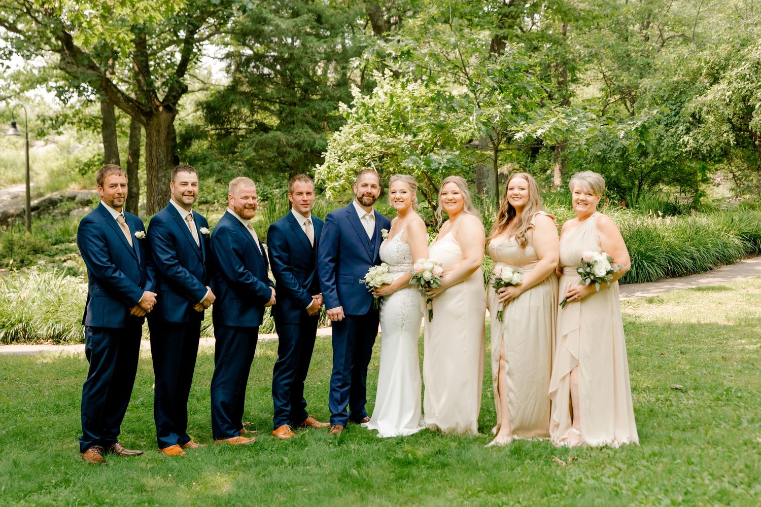 Wedding party of seven smiling outdoors with groomsmen in navy suits and bridesmaids in blush gowns.