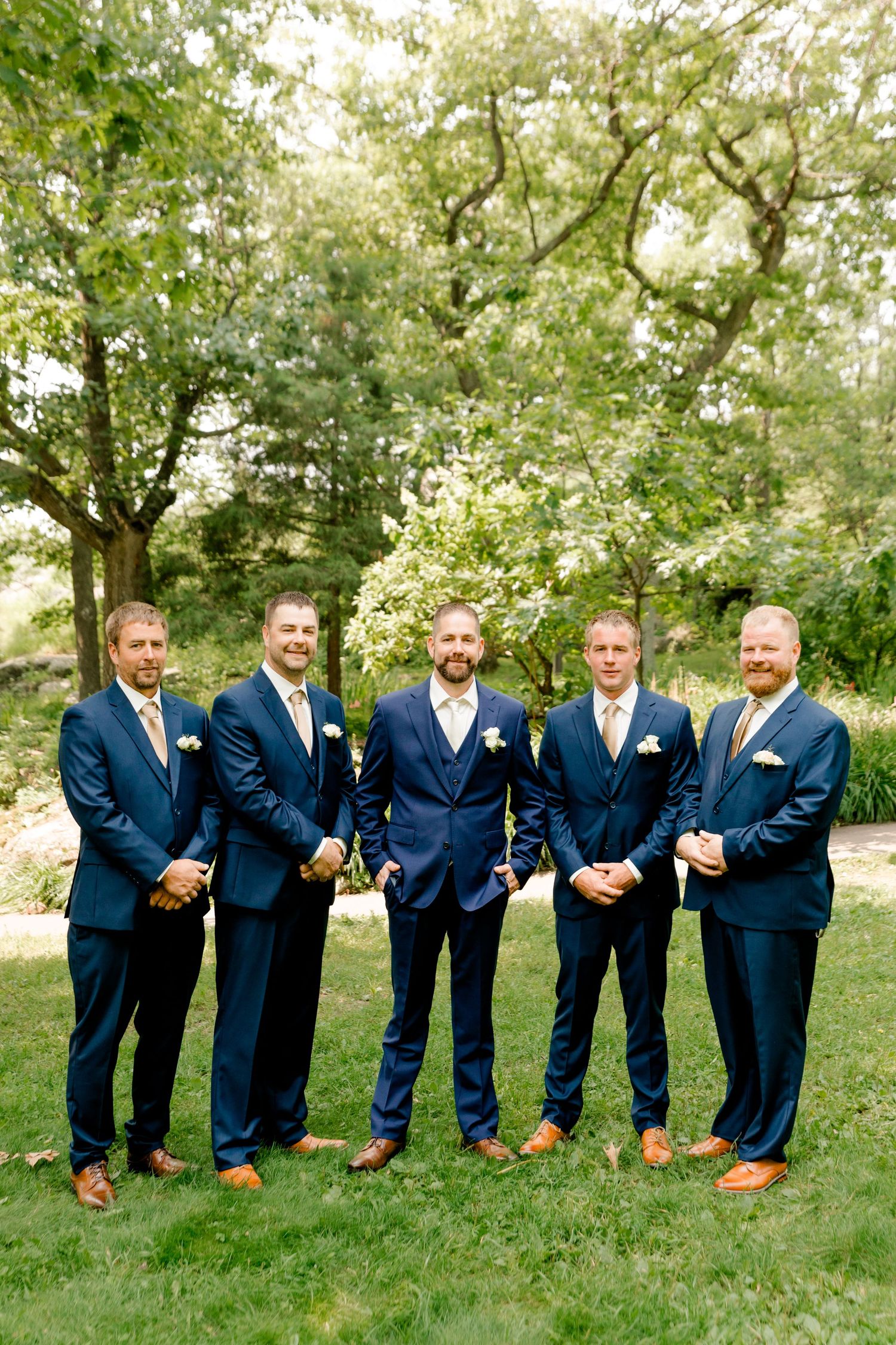 Five groomsmen in navy blue suits with gold ties pose outdoors on green grass for a wedding photo.