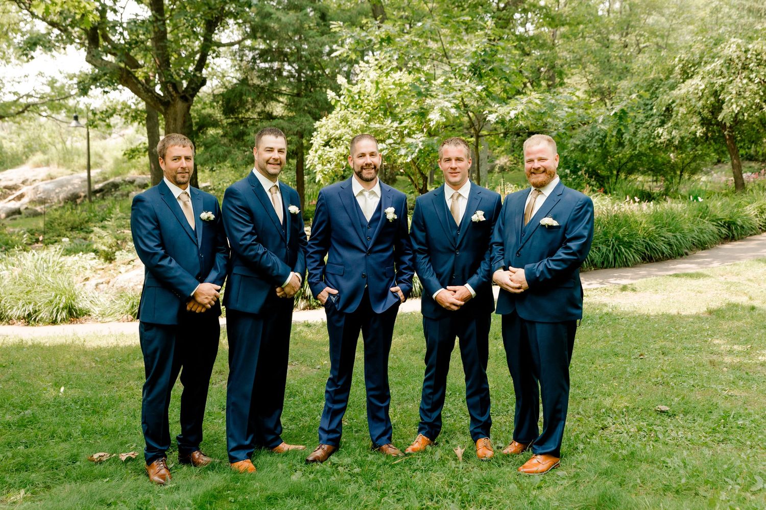 Five groomsmen in navy blue suits with brown shoes pose outdoors on green grass at a wedding ceremony.
