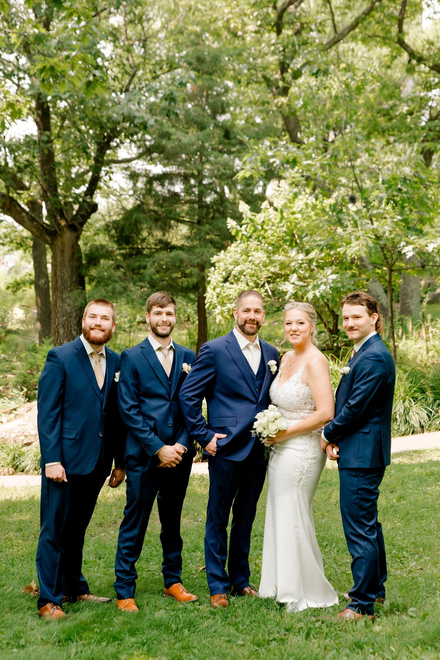 A bride in a white gown holds flowers beside her groom and three groomsmen in navy suits outdoors.
