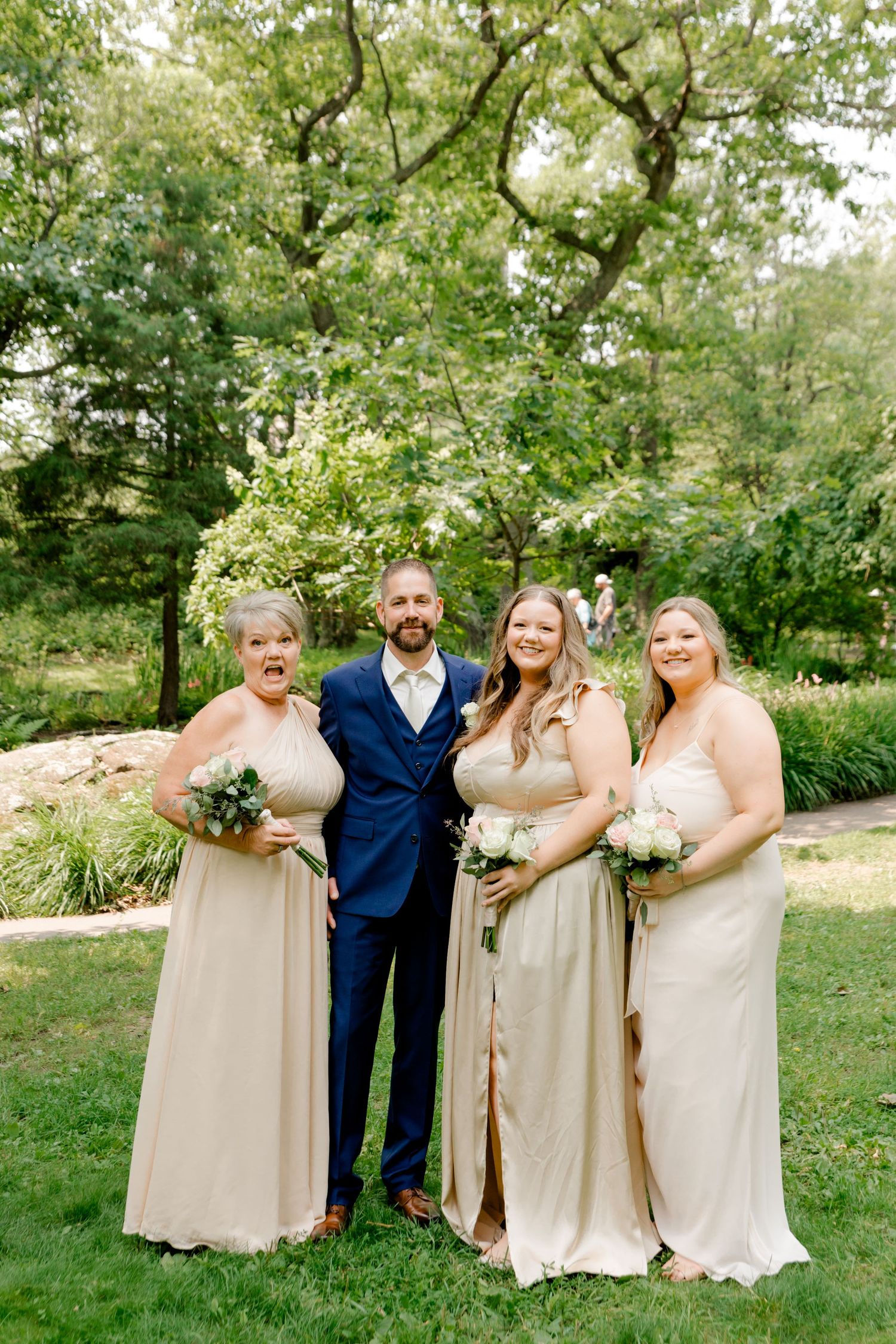 Groom in navy suit with three bridesmaids in champagne gowns holding white bouquets, posing outdoors in a lush garden setting.