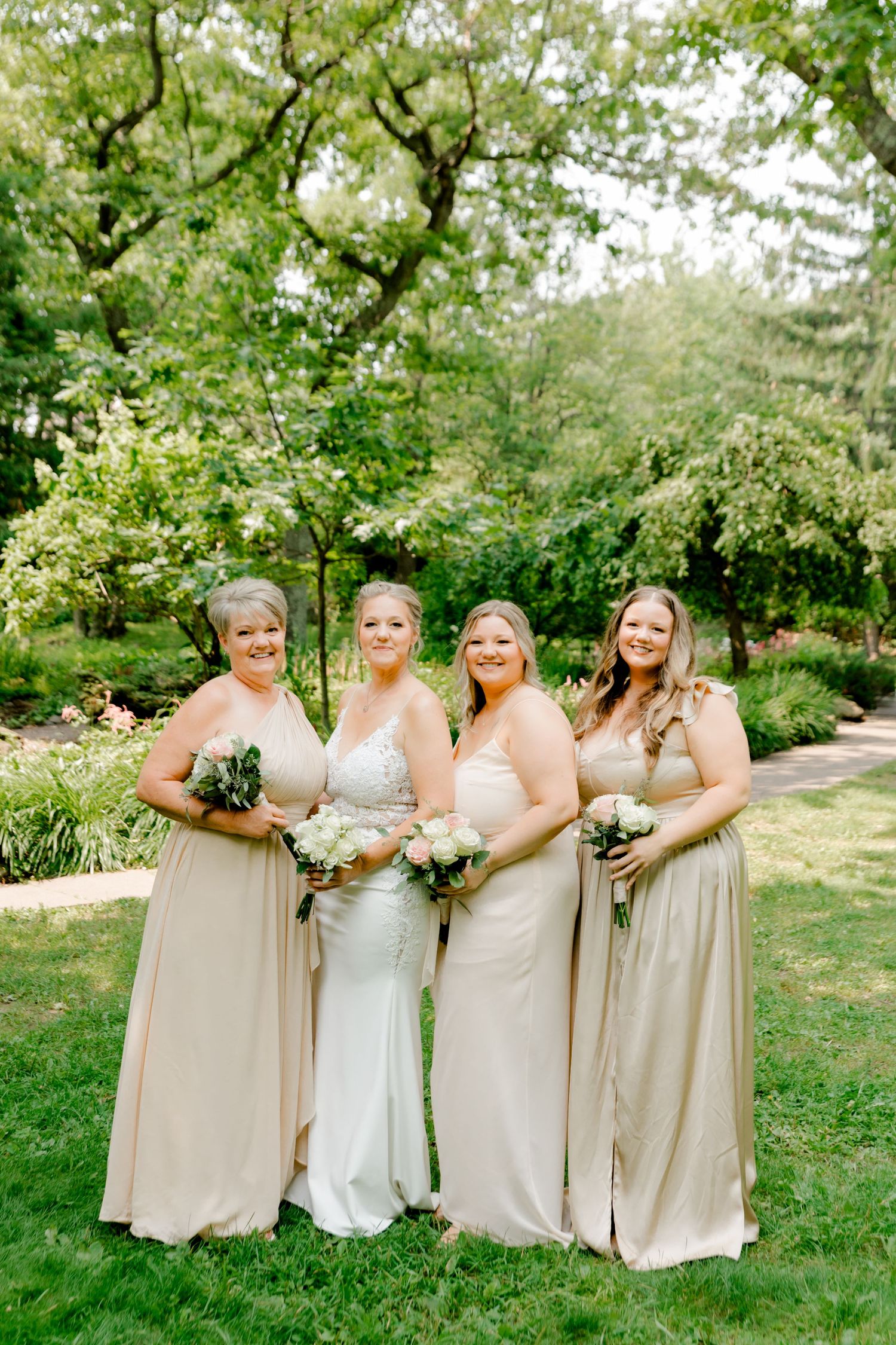 Bride and three bridesmaids in champagne gowns holding bouquets, posing outdoors in a lush green garden.