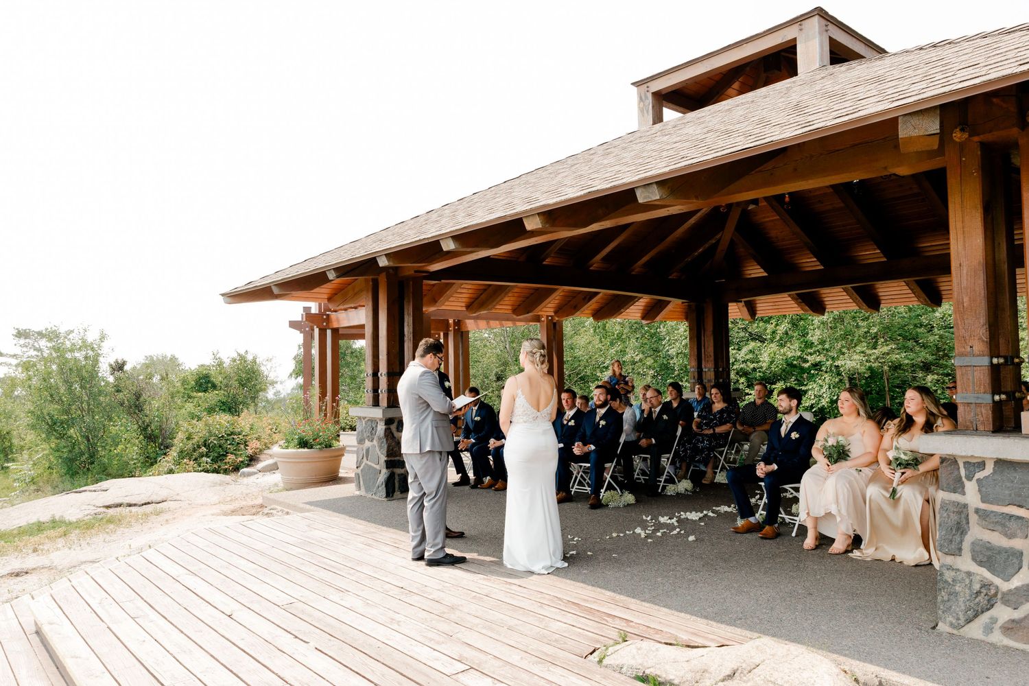 Outdoor wedding ceremony under a rustic wooden pavilion with guests seated, surrounded by lush greenery.