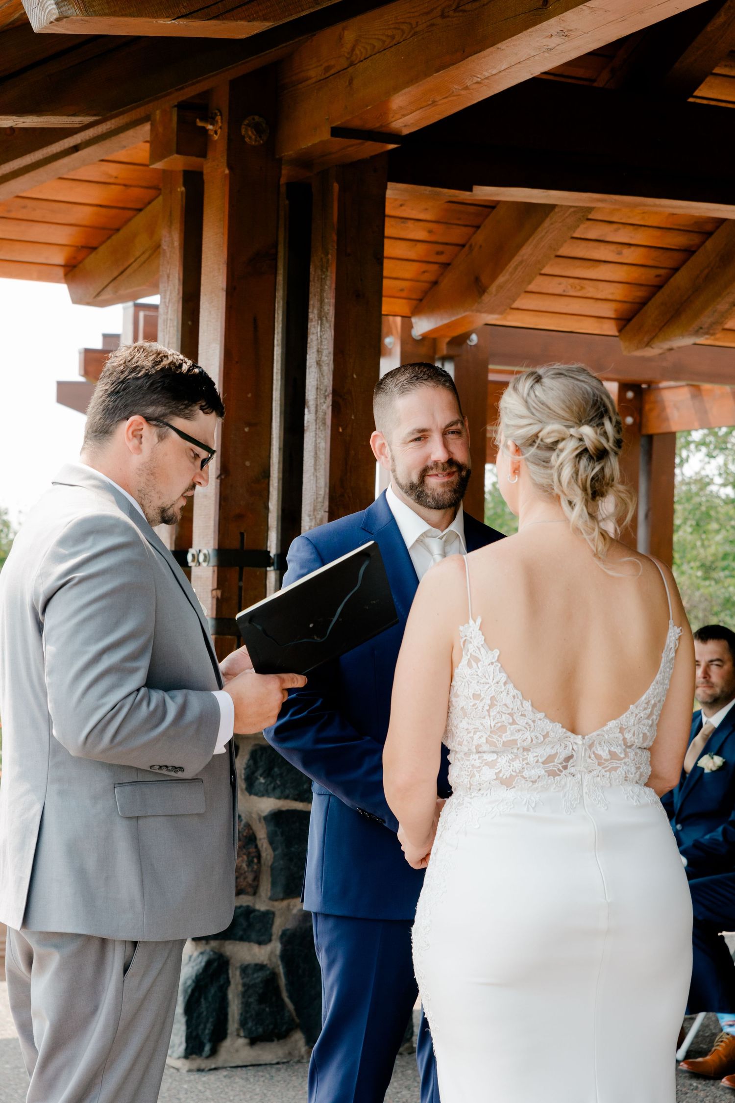 Bride and groom exchanging vows at outdoor wedding ceremony under wooden pavilion with officiant reading from black folder.