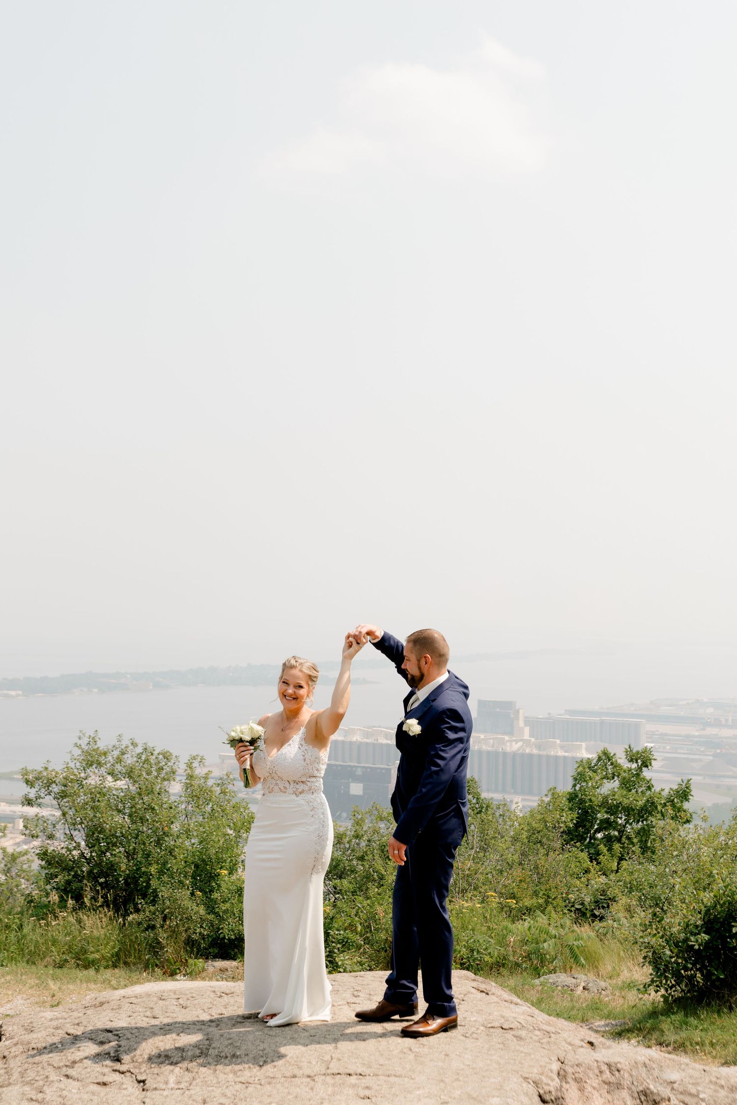 Bride and groom dancing on a rocky overlook with a scenic city and river view on their wedding day.