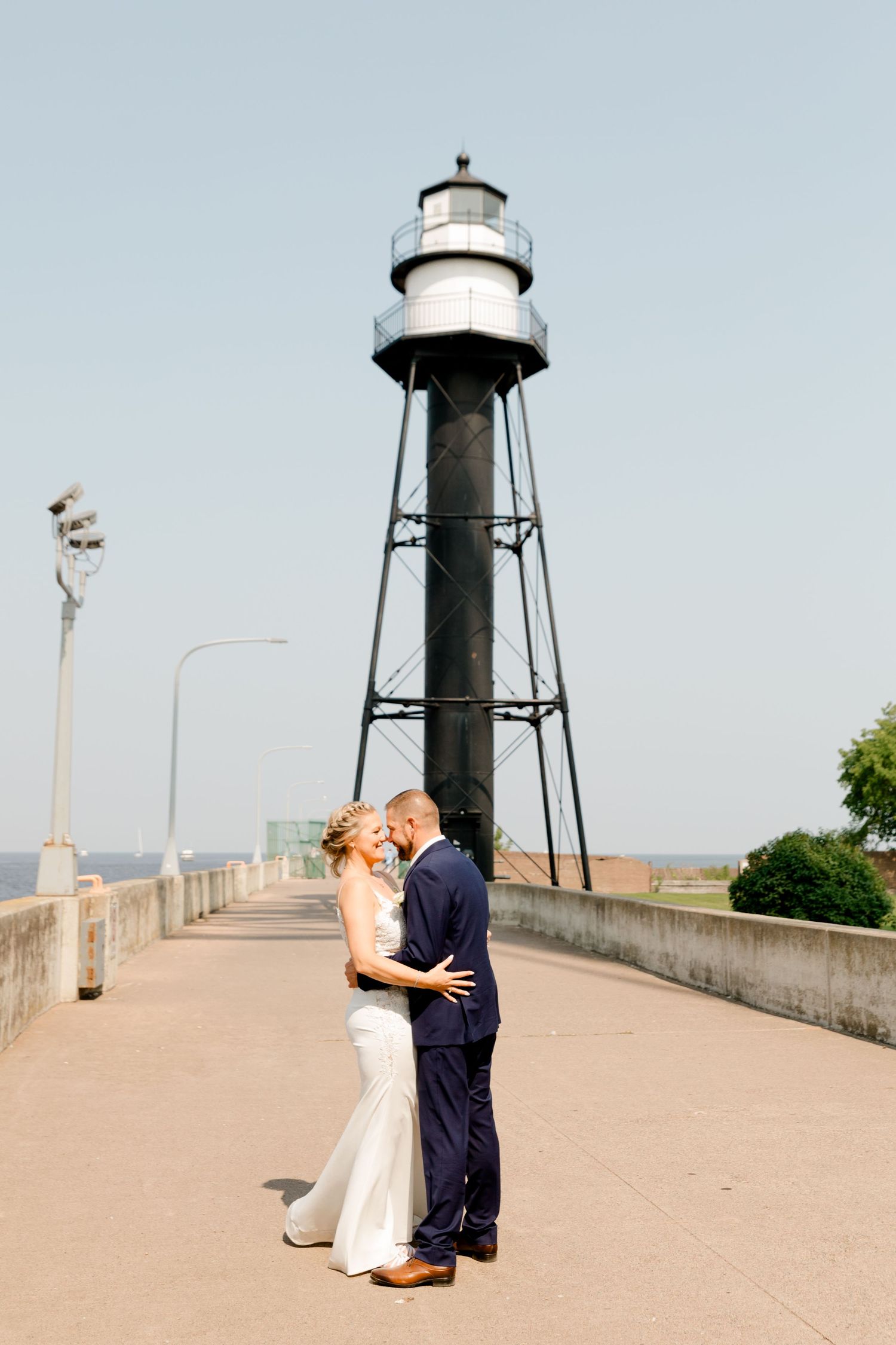 Bride and groom share a romantic moment in front of a tall lighthouse on a sunny wedding day.