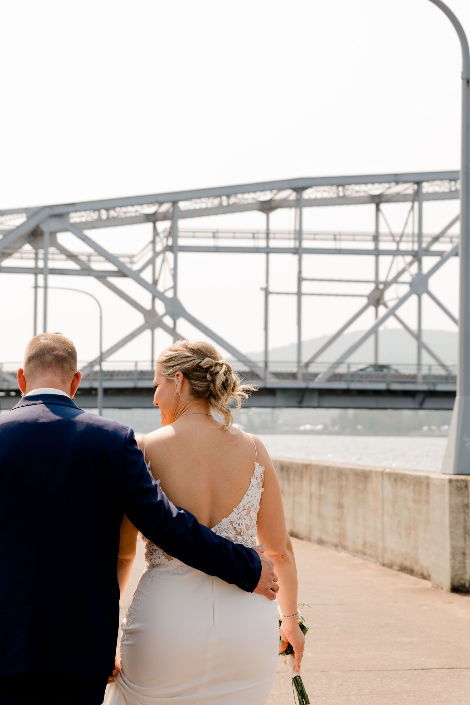 Bride and groom walk hand in hand along a waterfront promenade with a steel bridge backdrop.
