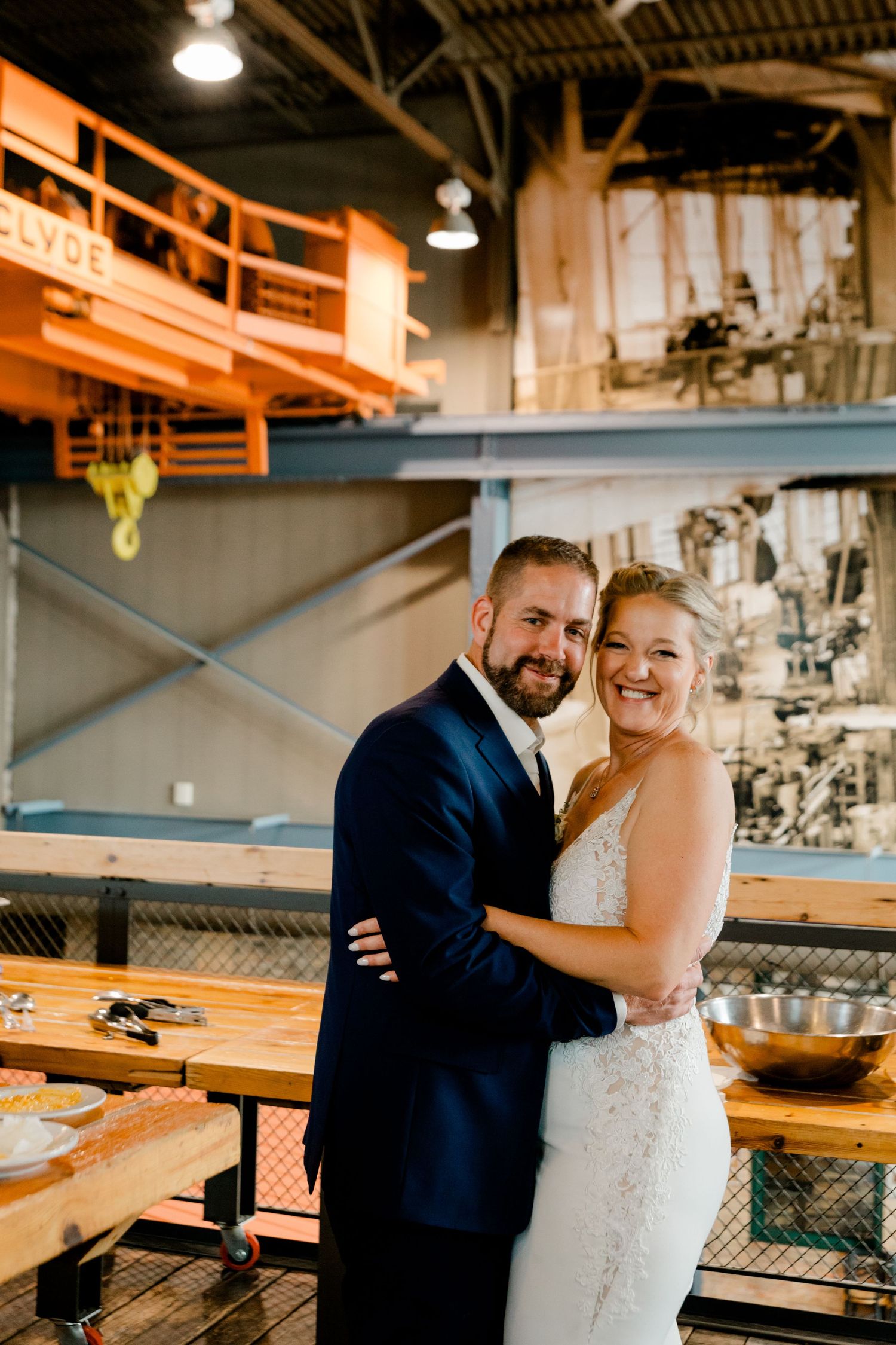 Happy bride and groom embrace inside a rustic industrial venue with orange crane and brick walls.