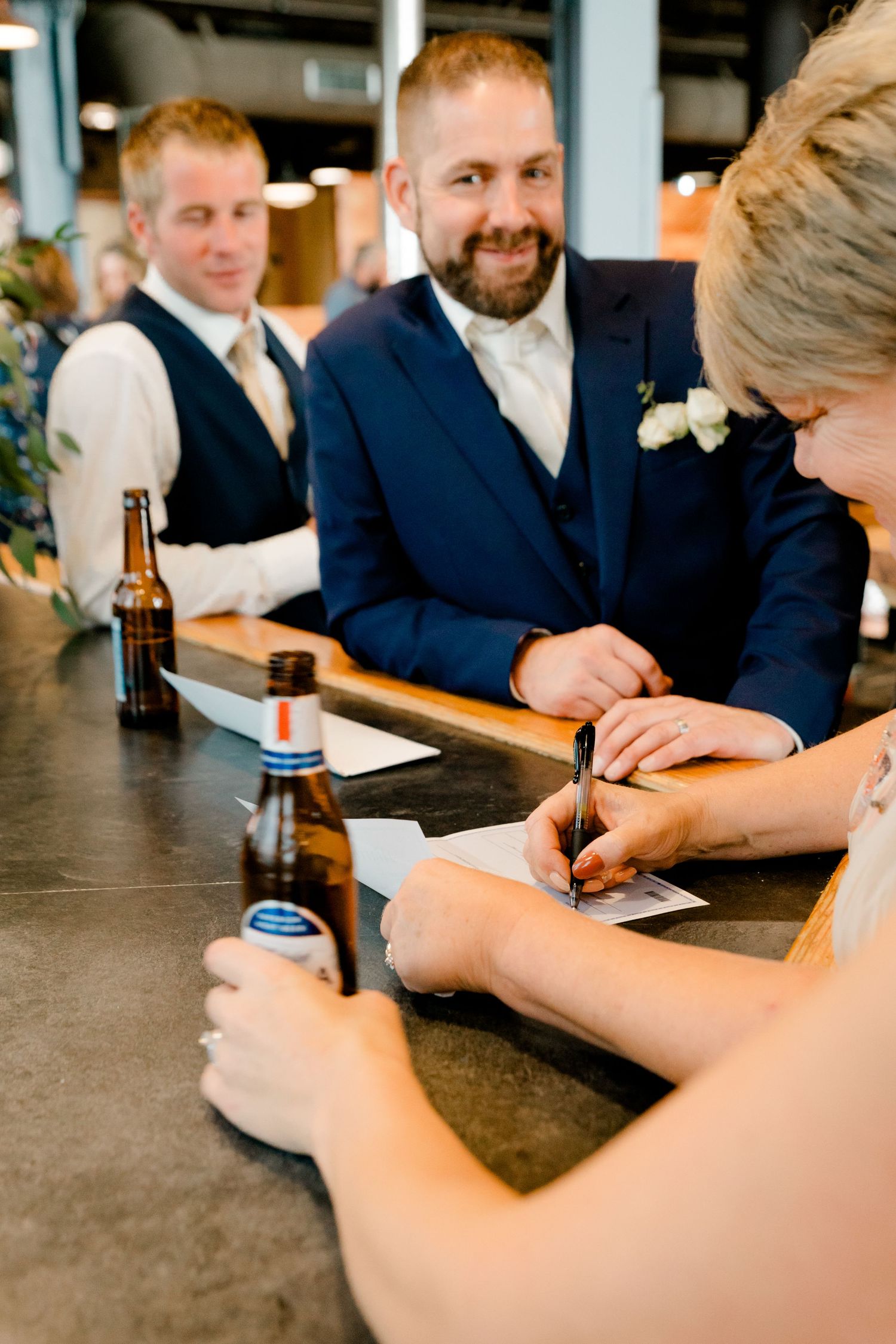 Groom in navy suit smiles as bride signs wedding documents at a bar counter with beer bottles nearby.