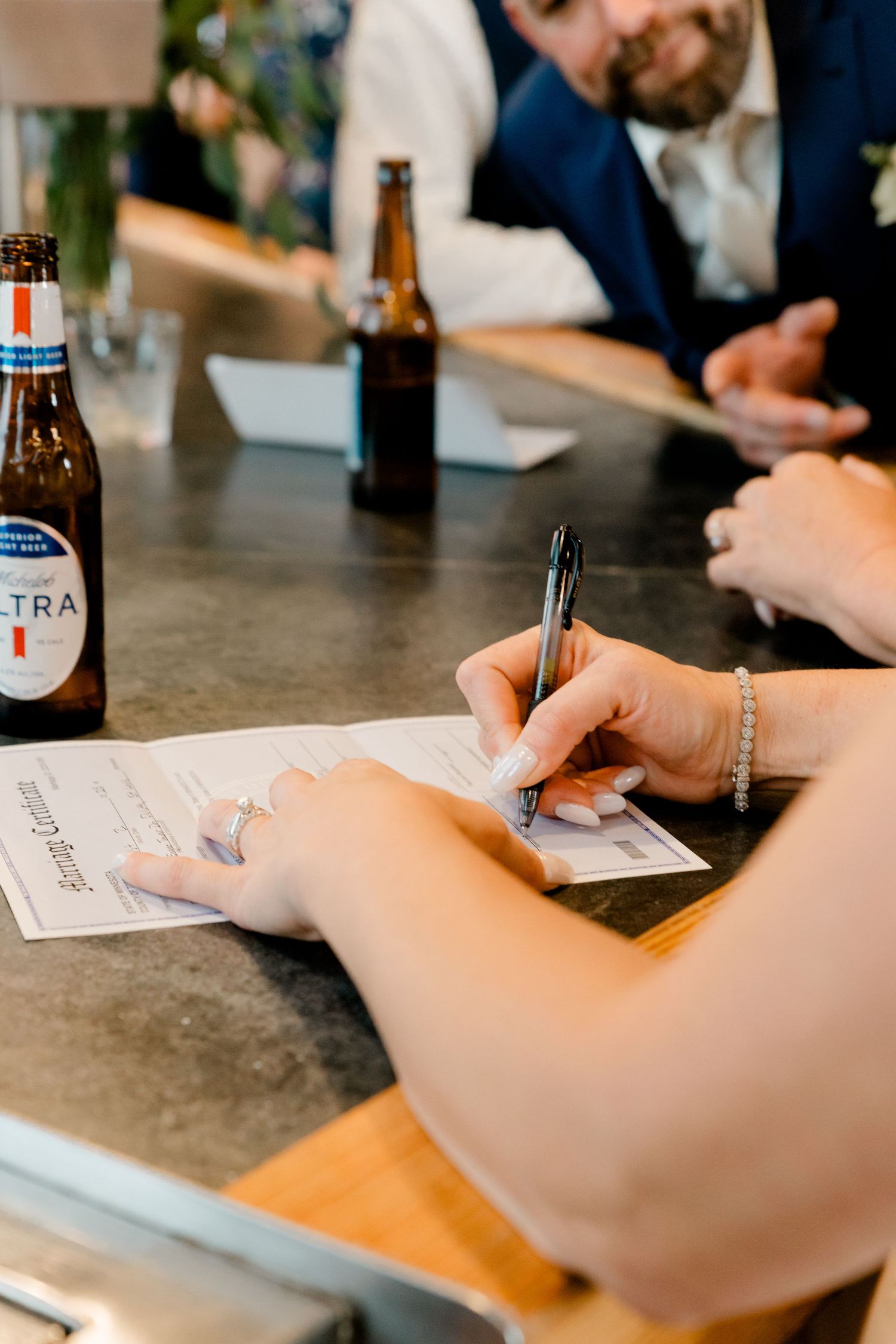 Close-up of bride signing wedding license at a bar counter with a Michelob Ultra bottle nearby.