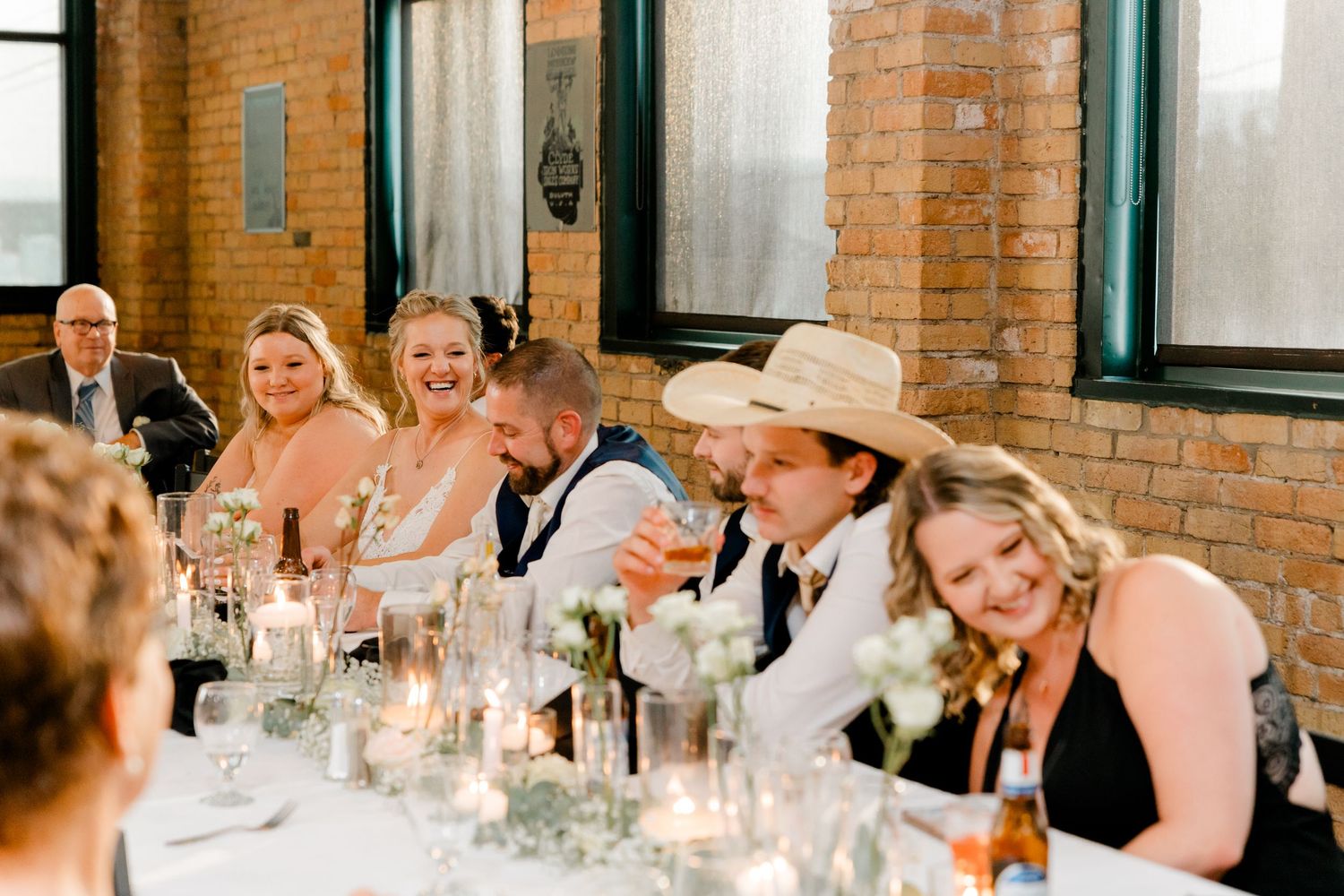 Wedding guests laugh and celebrate at a candlelit reception table inside a rustic brick venue.