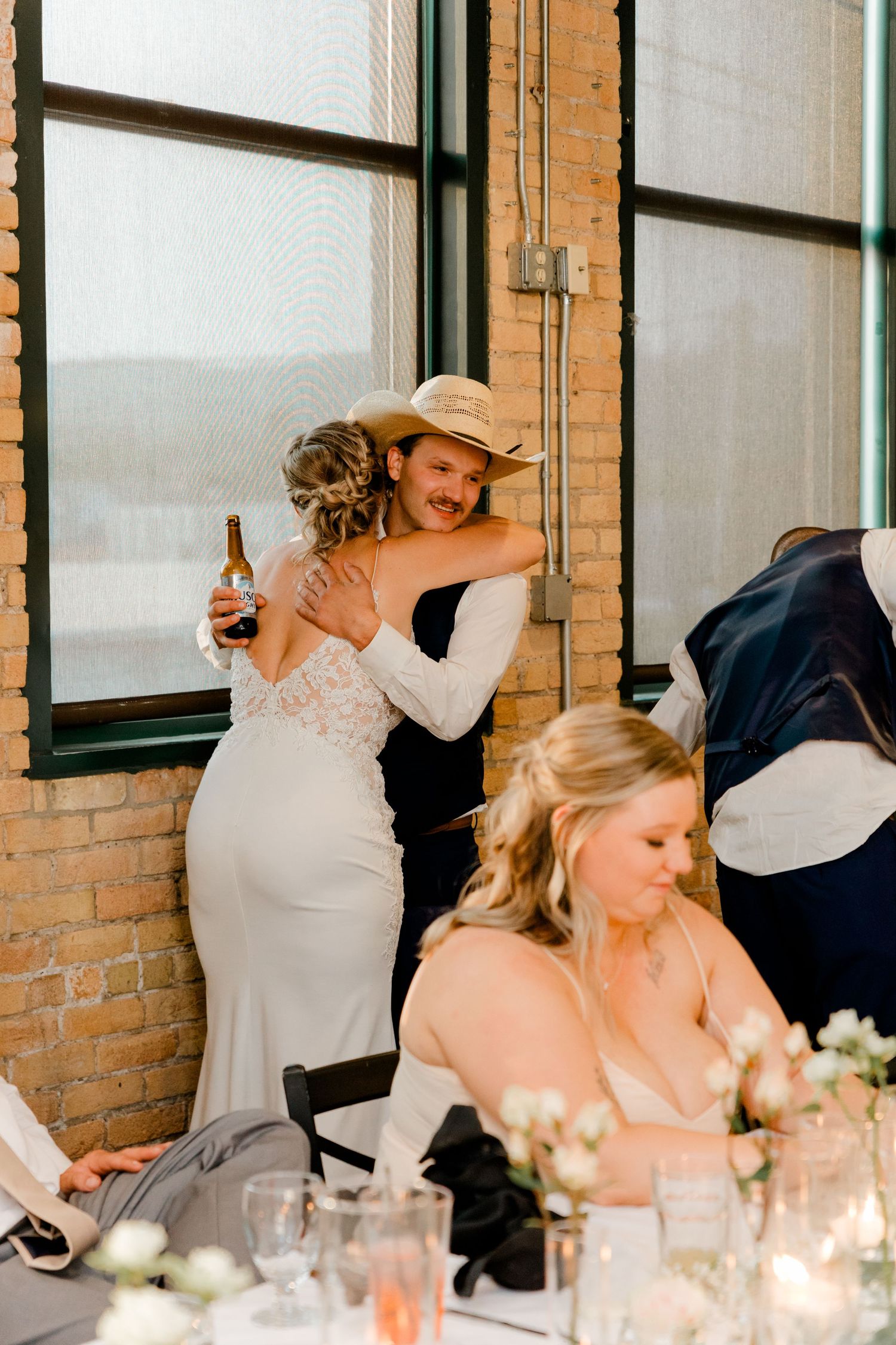 Bride in lace gown hugging guest in cowboy hat at elegant wedding reception with floral centerpieces.