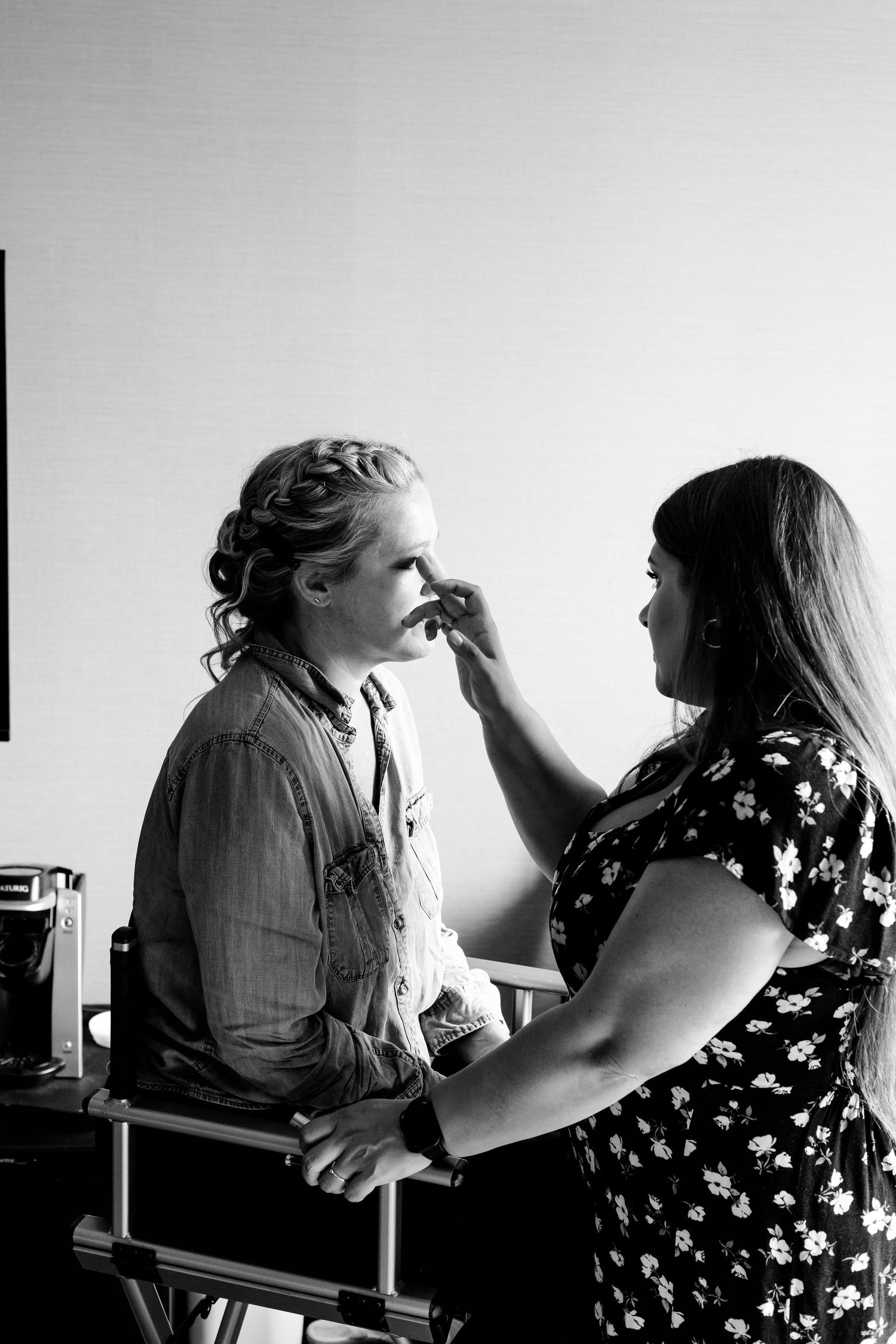 Black and white photo of a makeup artist applying makeup to a woman seated in a director's chair.