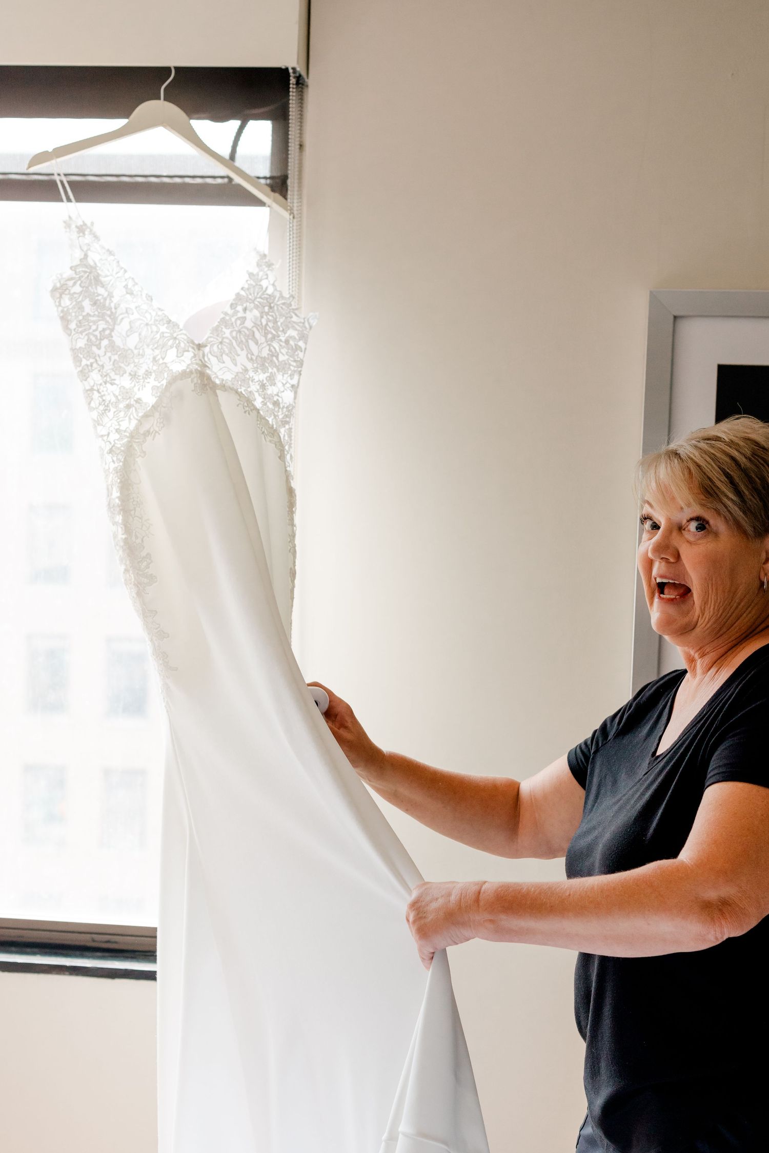 Woman reacting with surprise while holding a white beaded wedding dress hanging by a window.