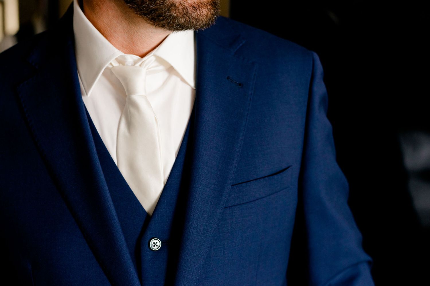 Man in navy blue three-piece suit with white dress shirt and ivory tie, elegant formal wedding attire.
