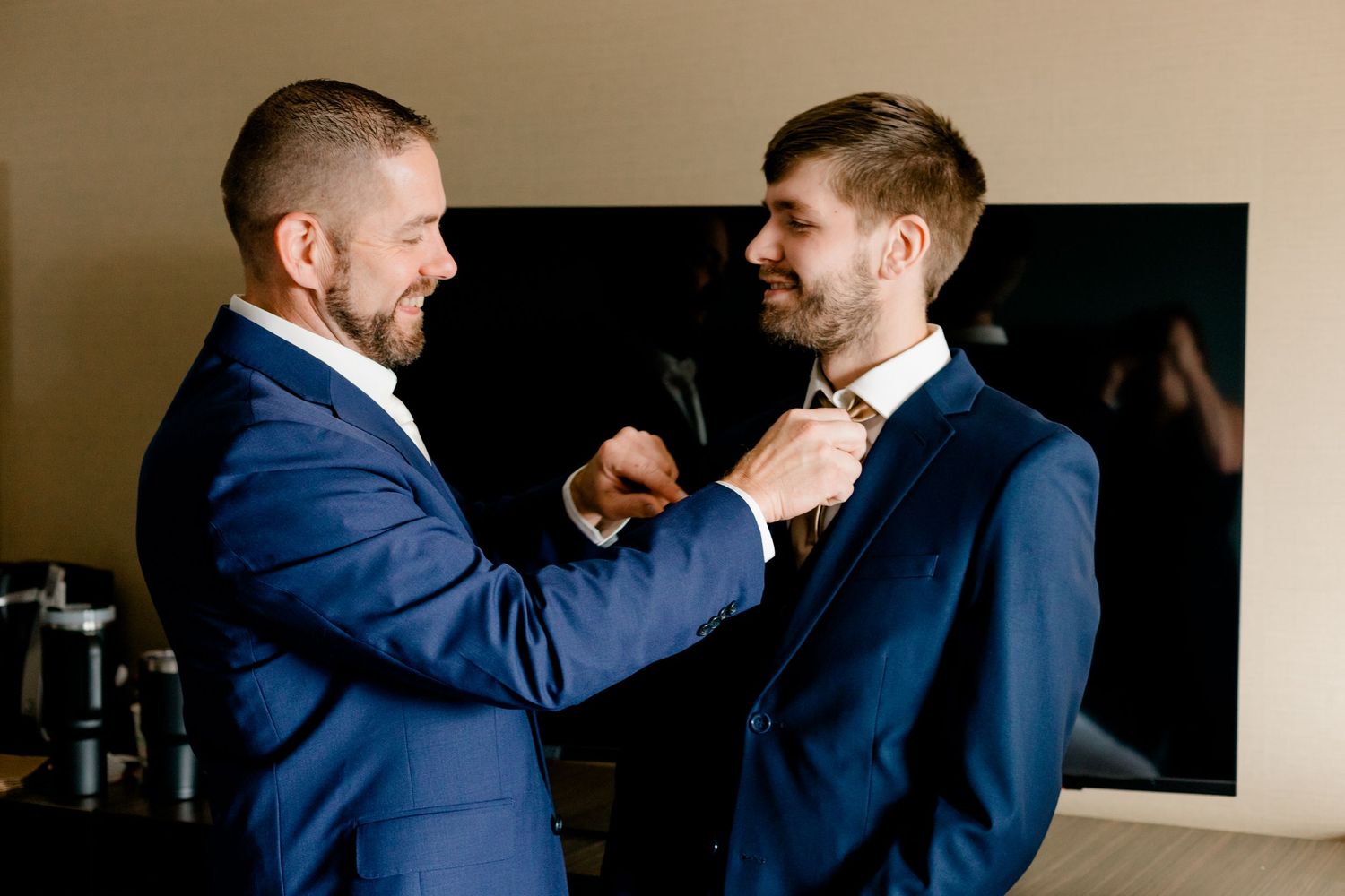 Two men in navy blue suits, one adjusting the other's bow tie while smiling, during wedding preparations.