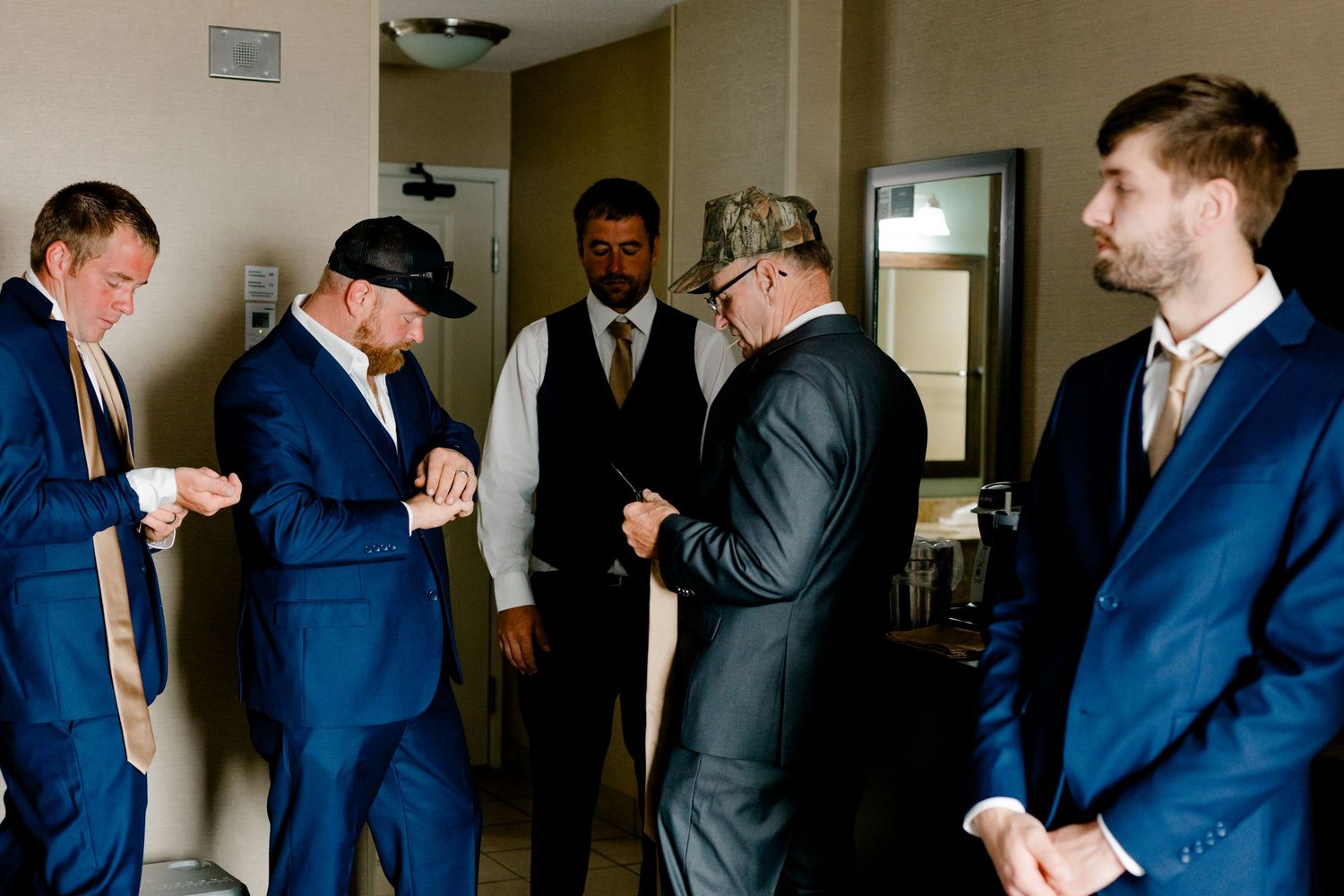 Groomsmen in navy suits help each other prepare before a wedding ceremony in a hotel room.