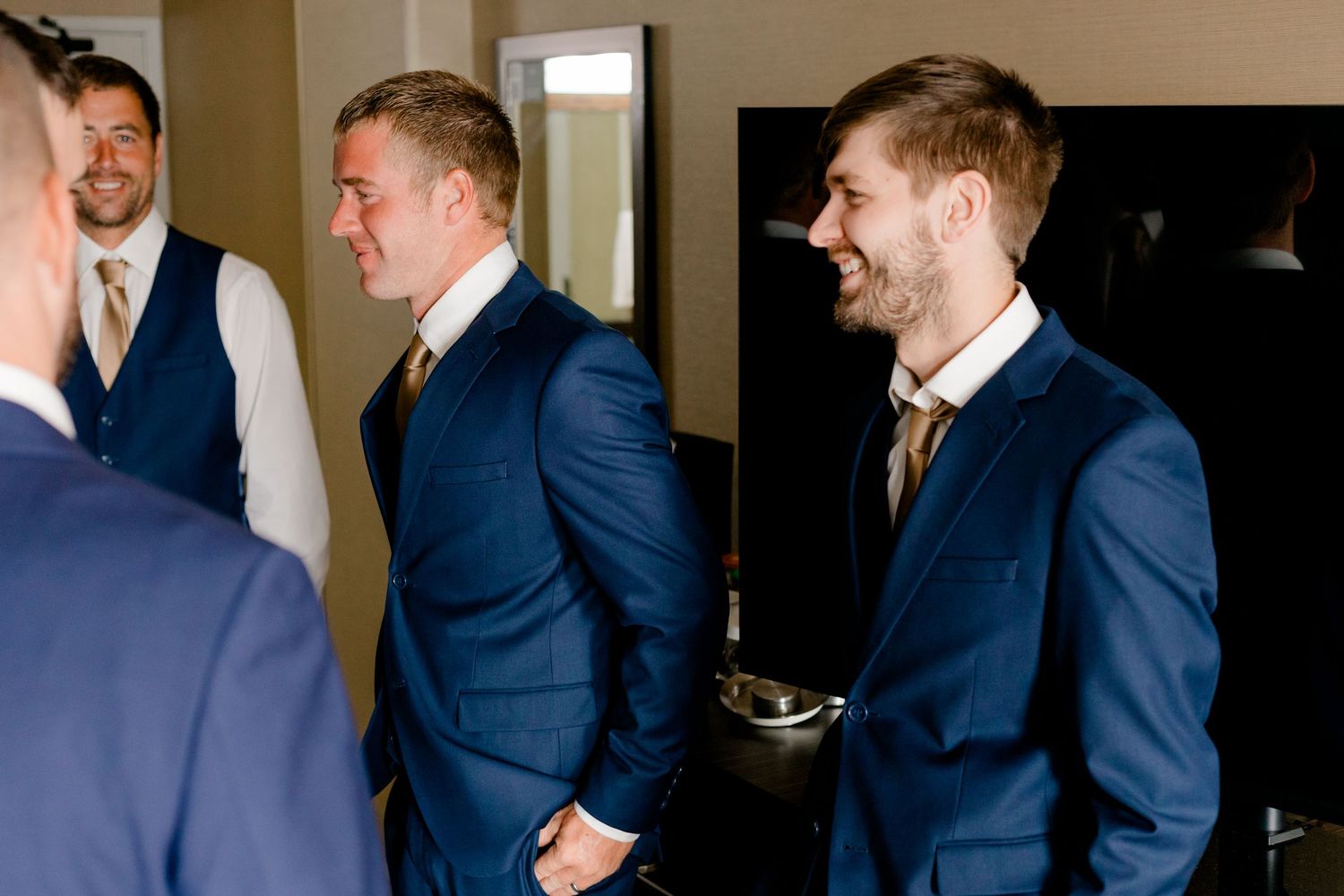 Groomsmen in navy blue suits with gold ties laughing and getting ready together in a hotel room before a wedding.