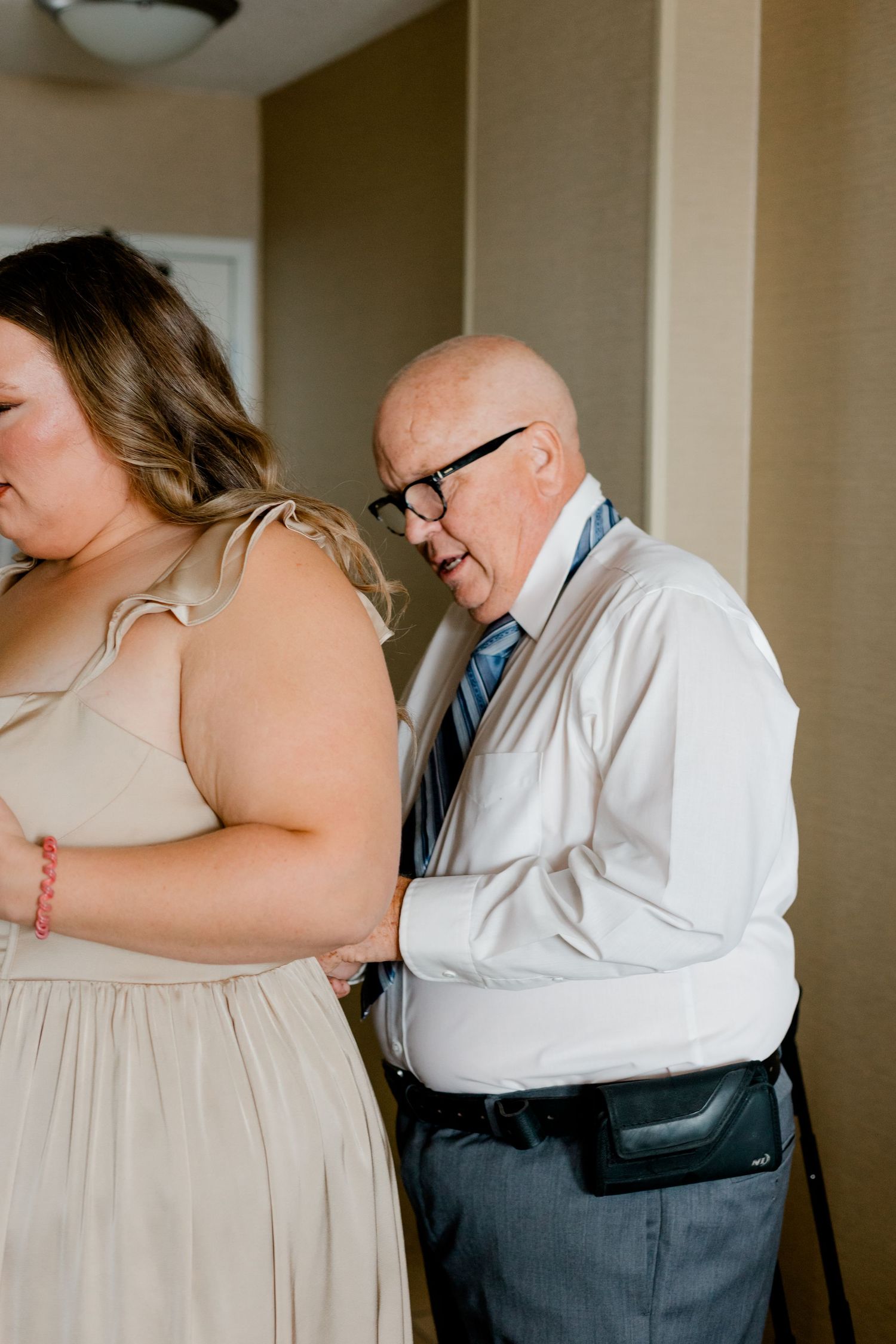 Elderly man in white shirt and tie helping zip up a beige bridesmaid dress at a wedding.