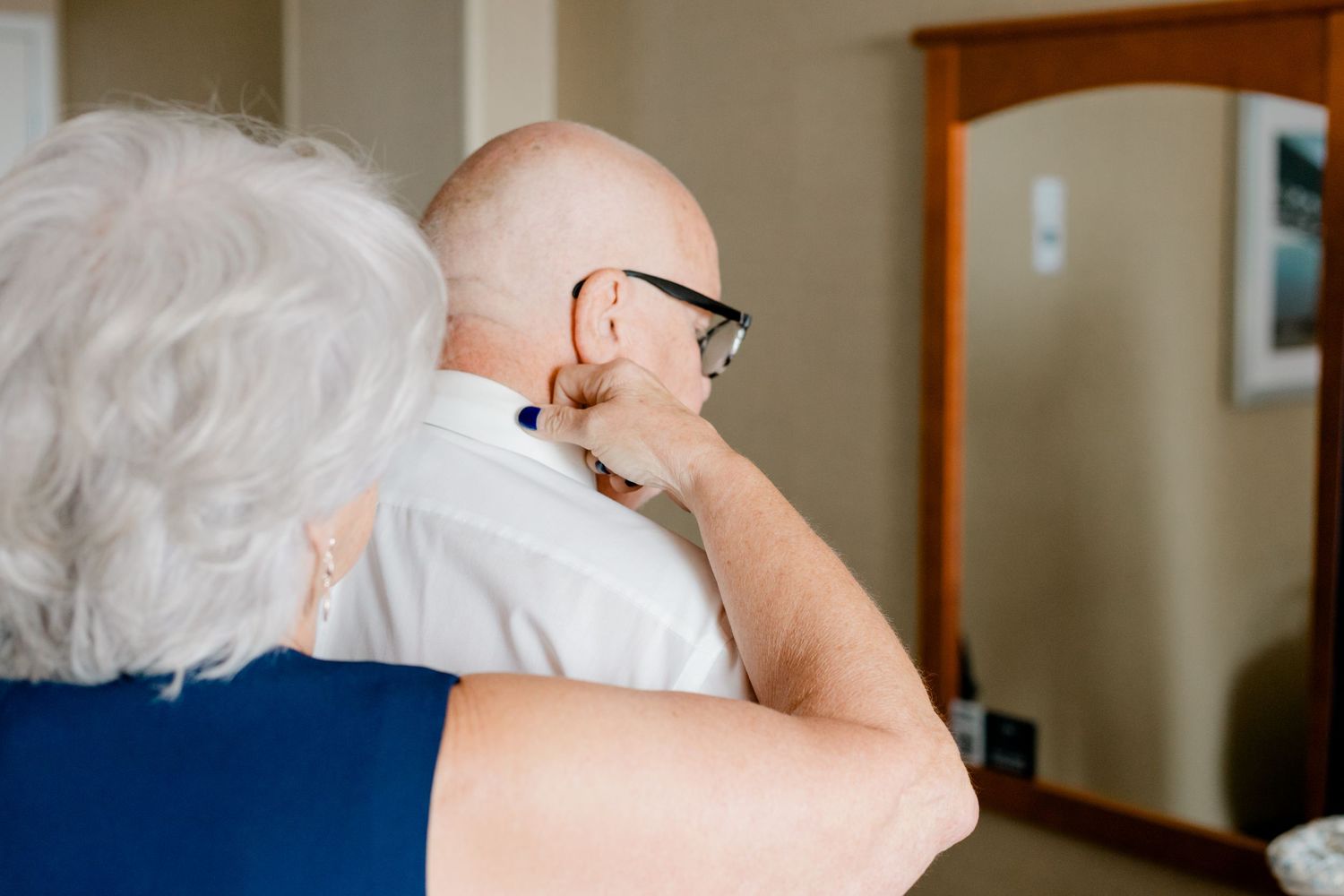 Elderly couple embracing from behind, bald man with glasses adjusting tie near mirror in bright room.