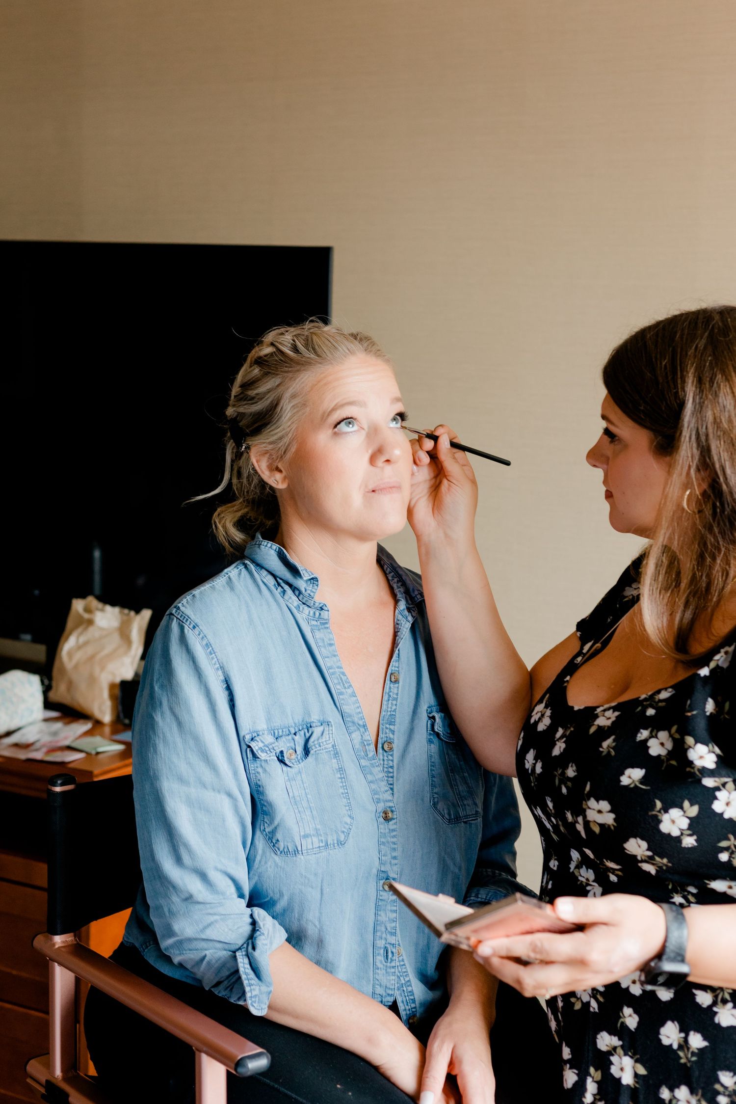 Makeup artist applying eye makeup to a blonde bride in a denim shirt during wedding preparations.