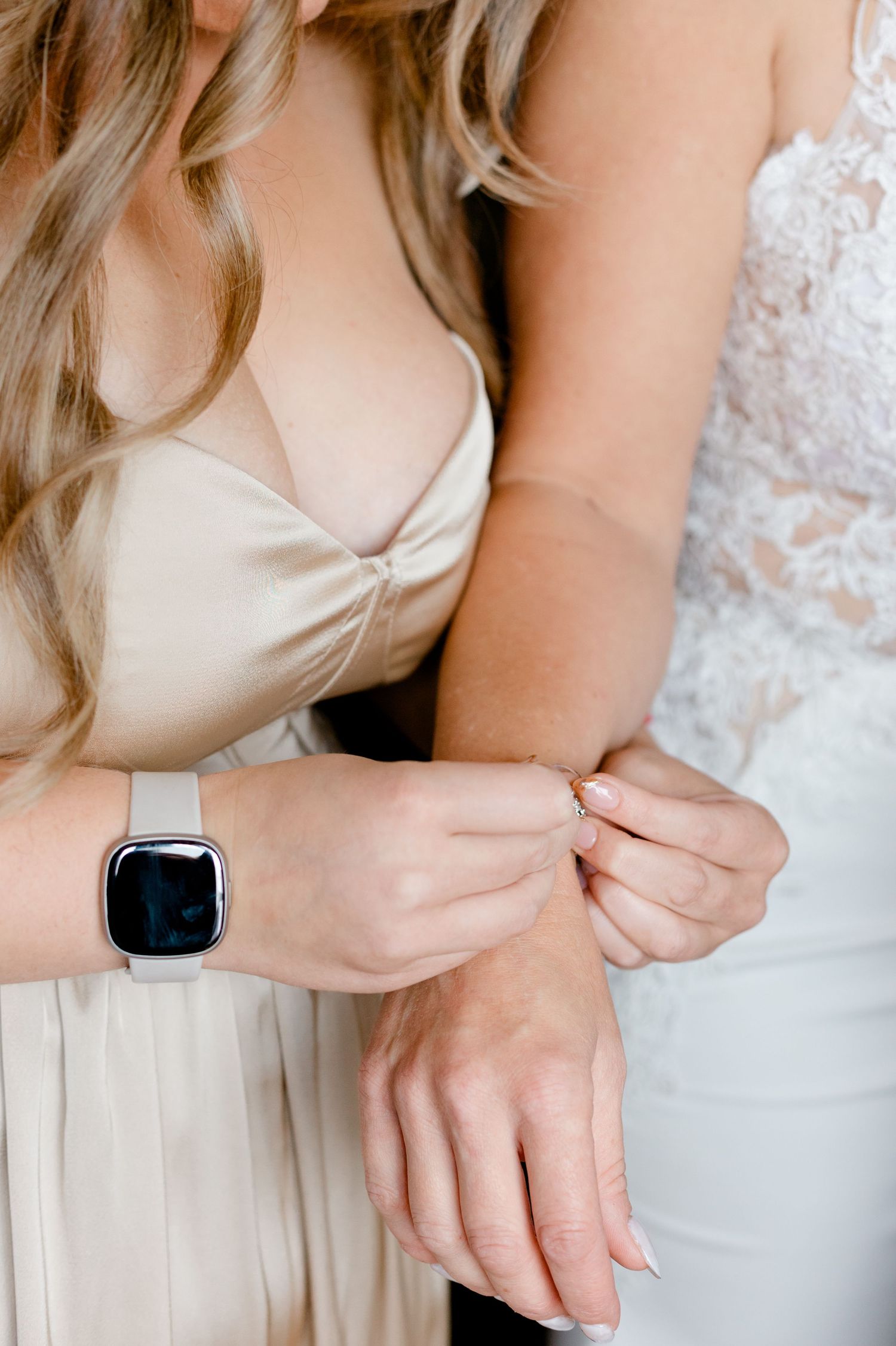 Bridesmaid in champagne satin dress helping bride put on a delicate gold bracelet on her wrist.