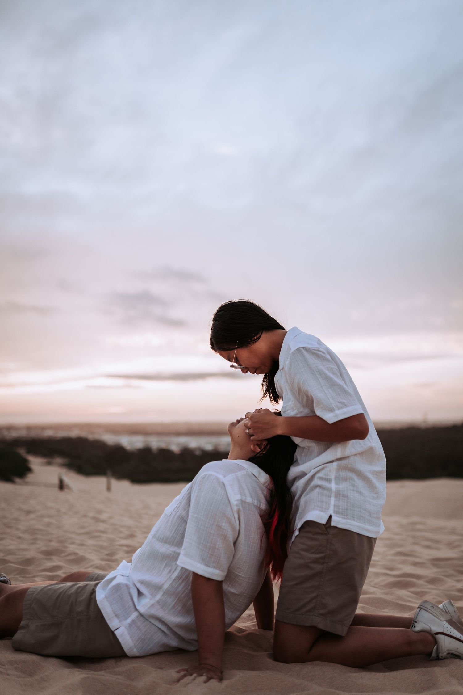 Two people in white shirts share an intimate moment on sand dunes during sunset with cloudy sky in background.