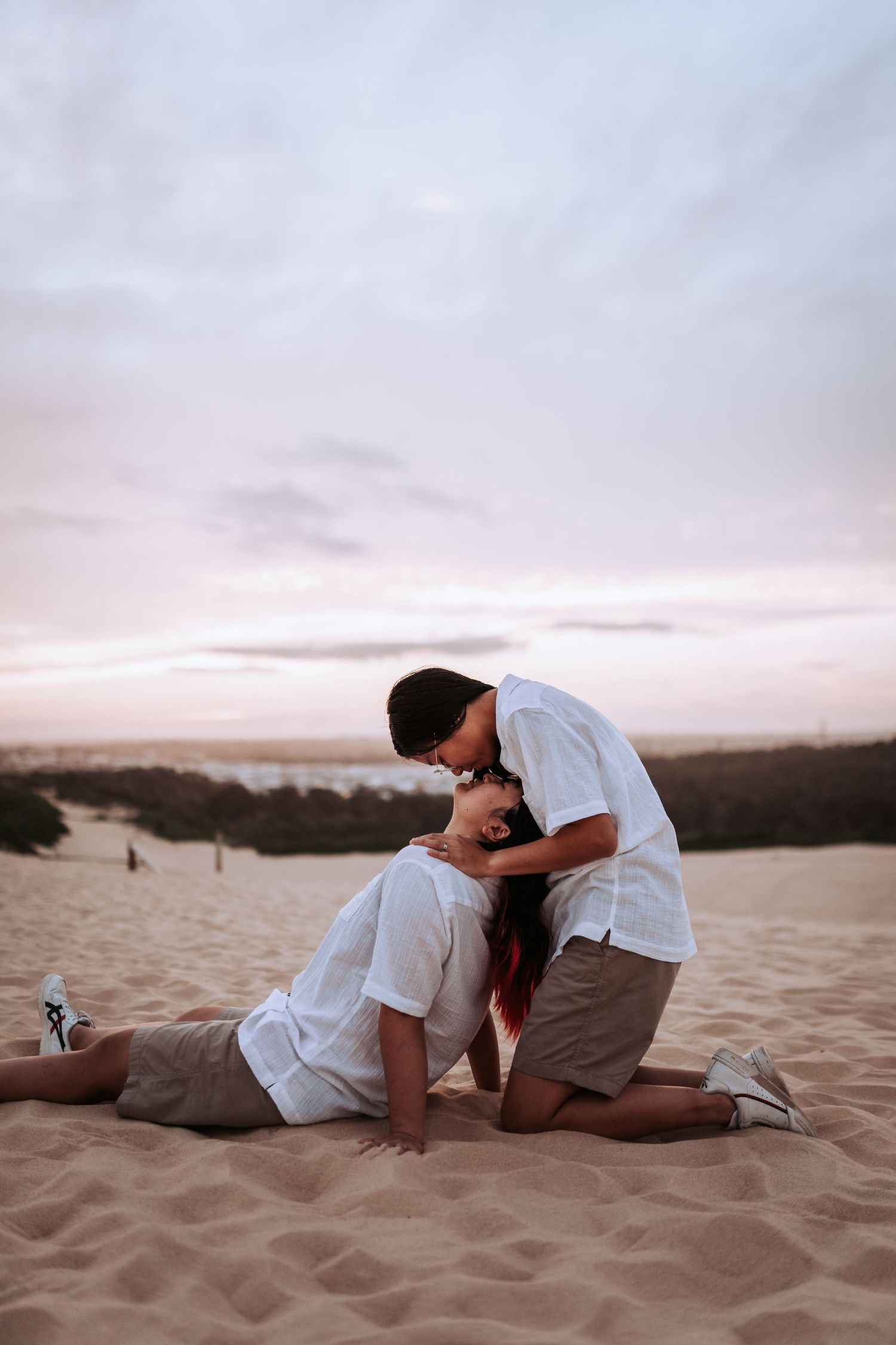 A romantic couple in white shares a tender moment on sandy dunes at sunset with ocean views in the background.