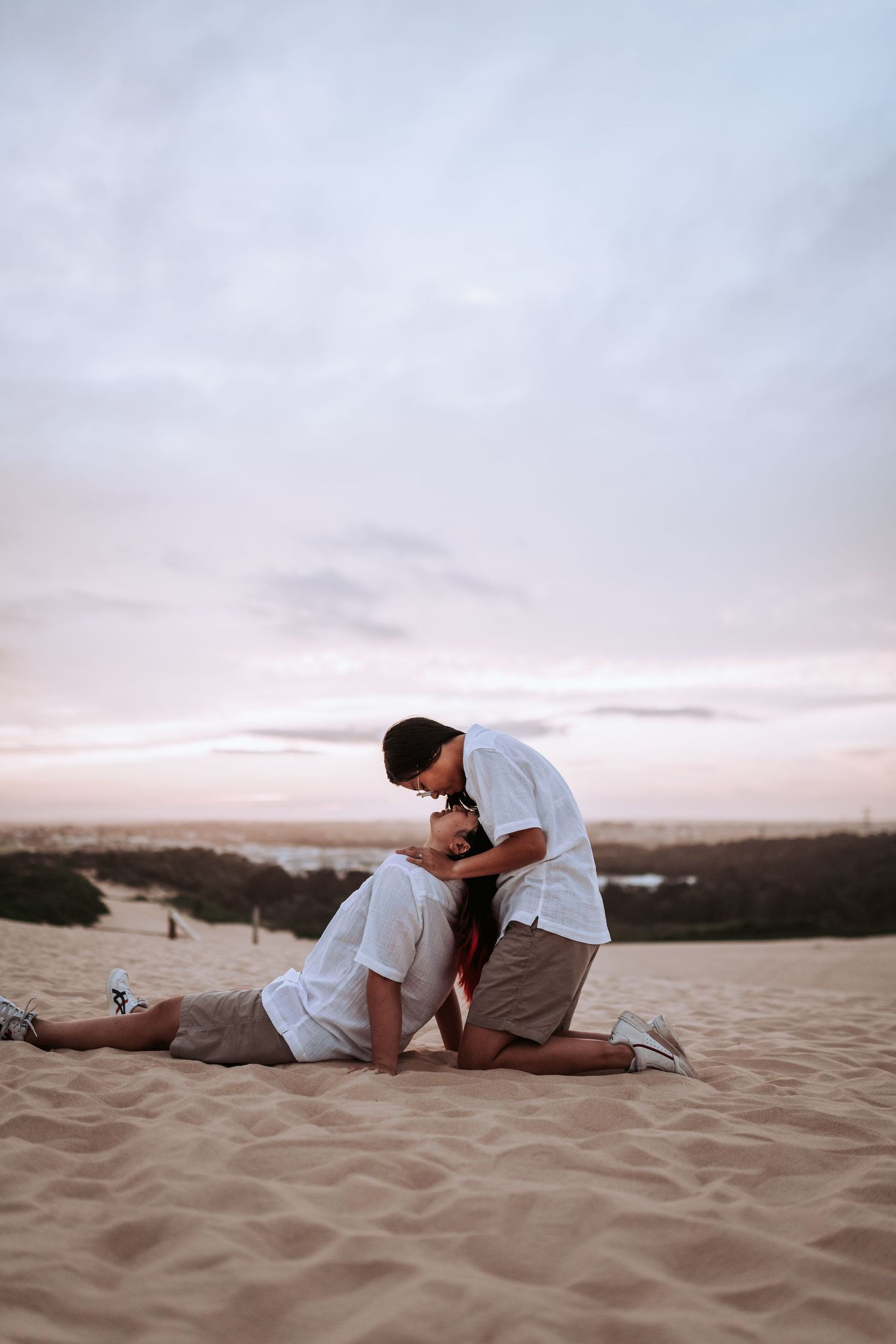 A romantic couple sharing an intimate moment while kneeling on sandy dunes at sunset.