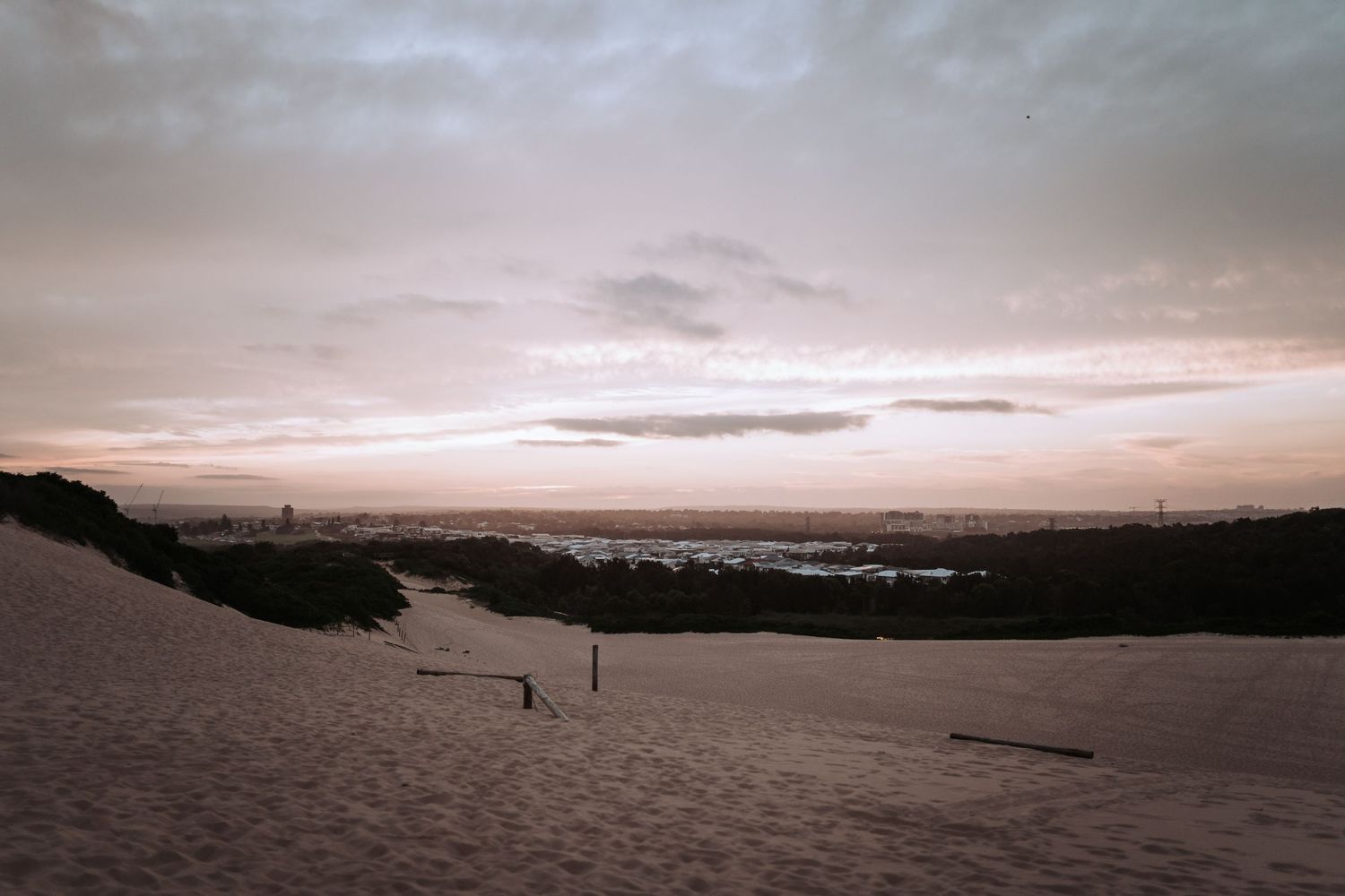 A sandy dune overlooks a coastal landscape at sunset with pinkish clouds in the sky.