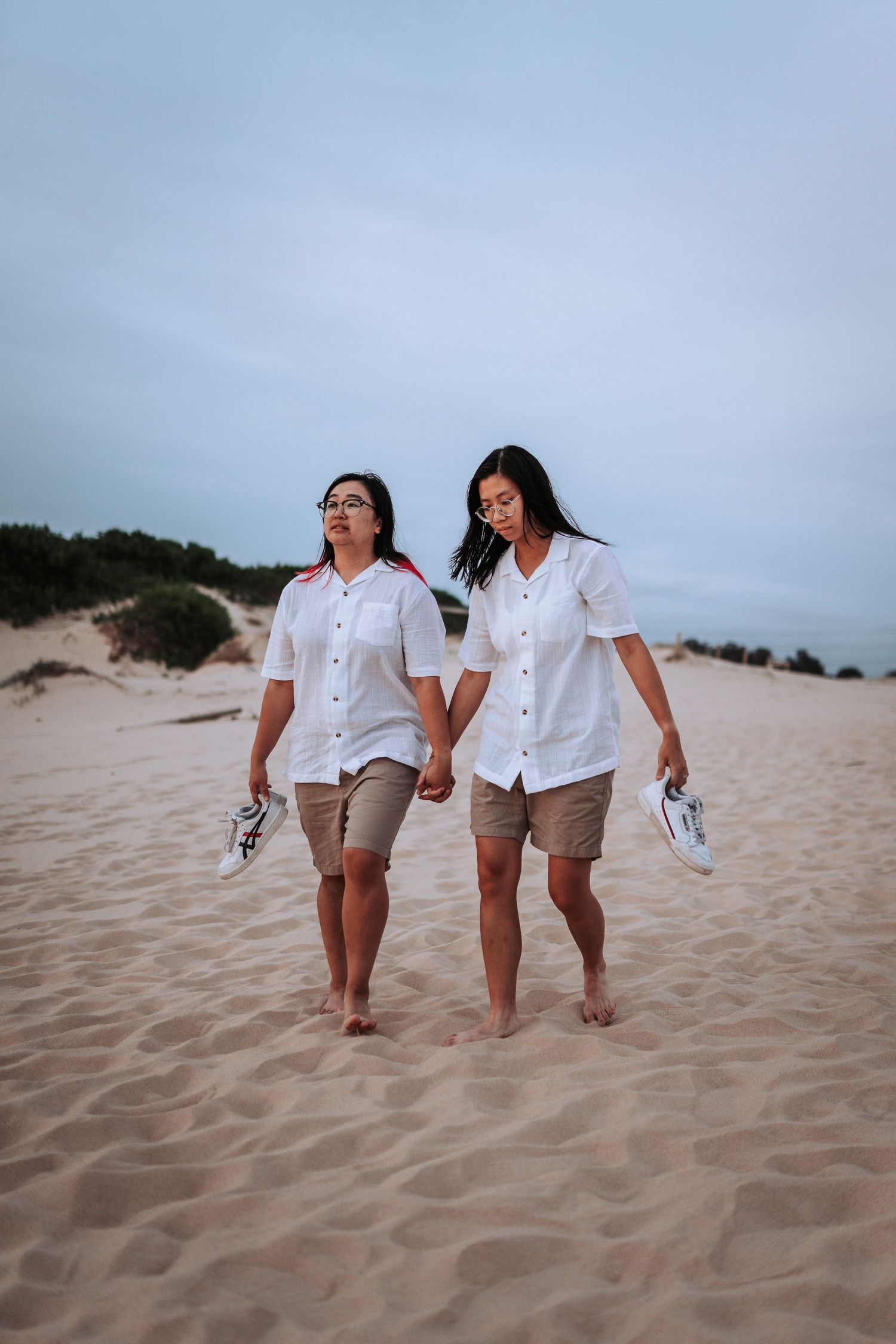 Two people walking hand in hand along a scenic beach dune wearing matching white shirts and khaki shorts.