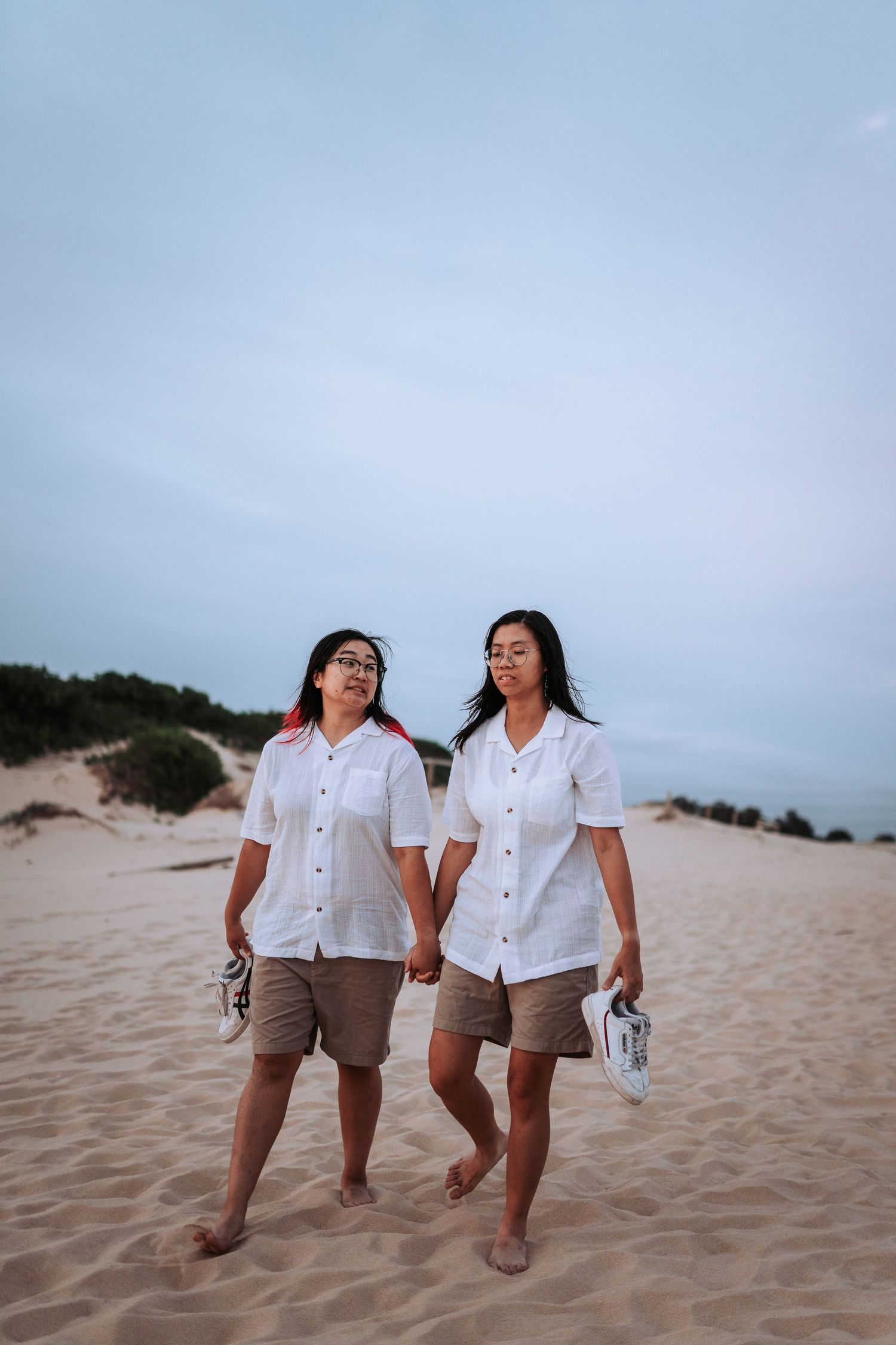 Two people in matching white shirts and khaki shorts walk together holding hands while barefoot on sandy dunes.