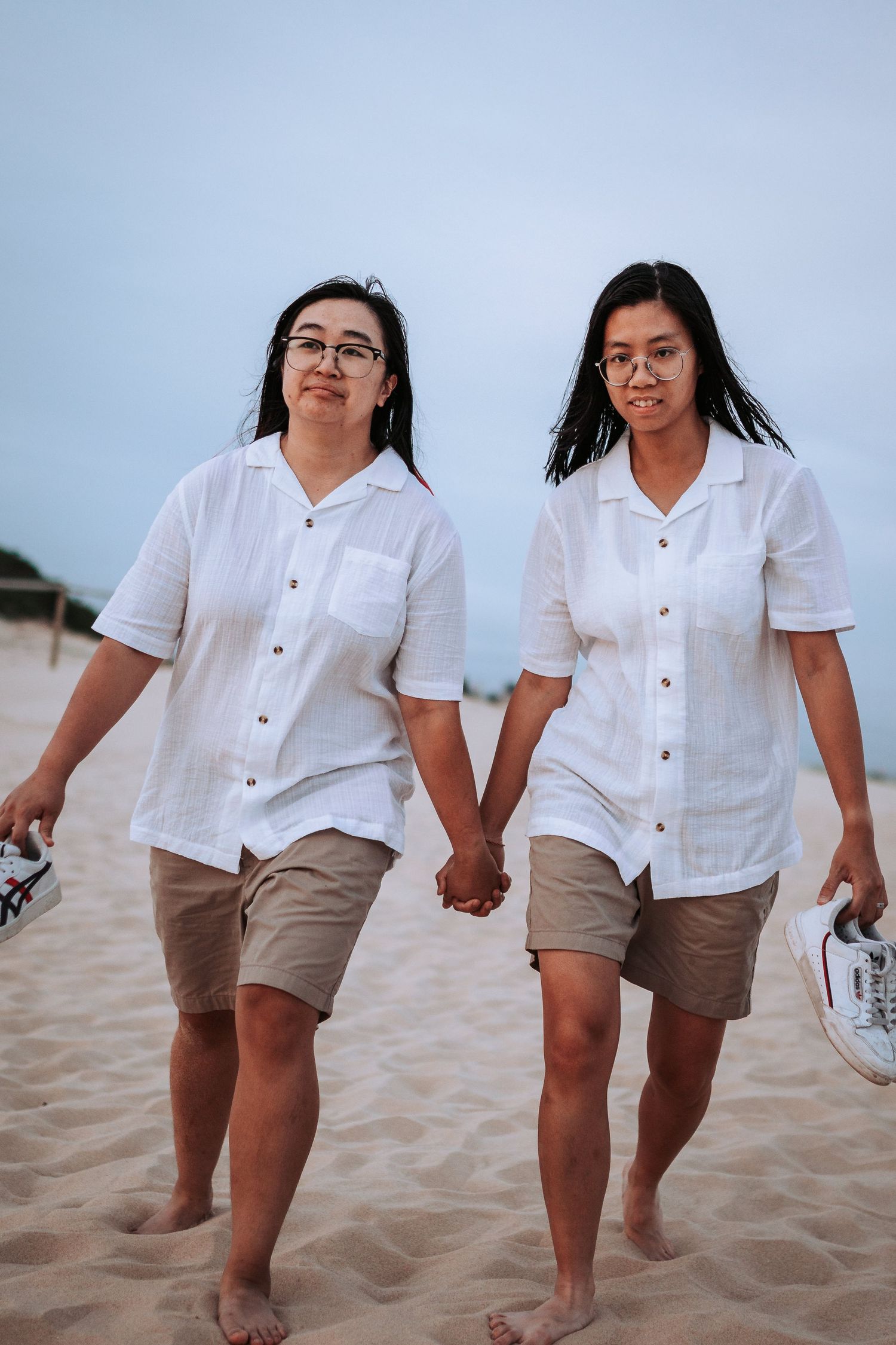 Two people walking hand in hand on a sandy beach wearing matching white button-up shirts and brown shorts.