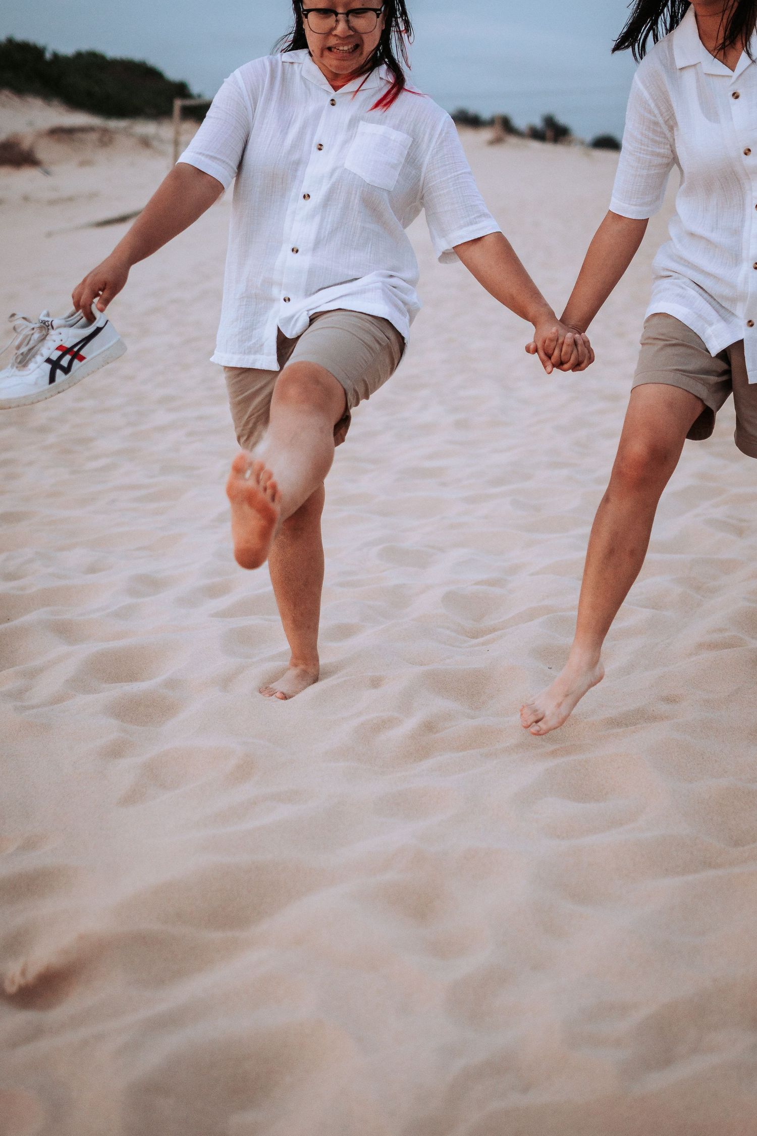 Two people in white shirts and khaki shorts walking barefoot on soft sand at the beach.