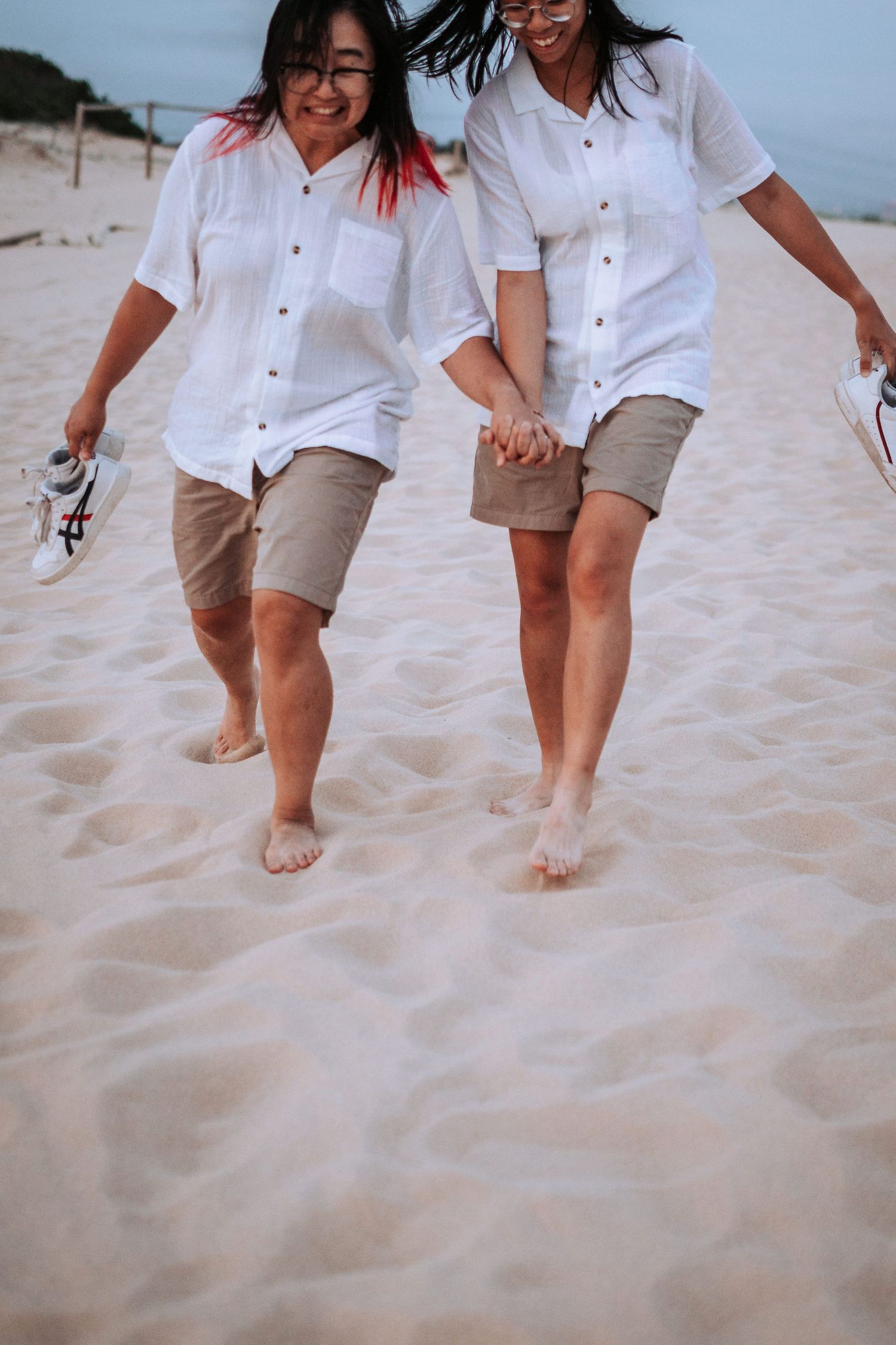 Two people in matching white shirts and khaki shorts walking barefoot together on a sandy beach.