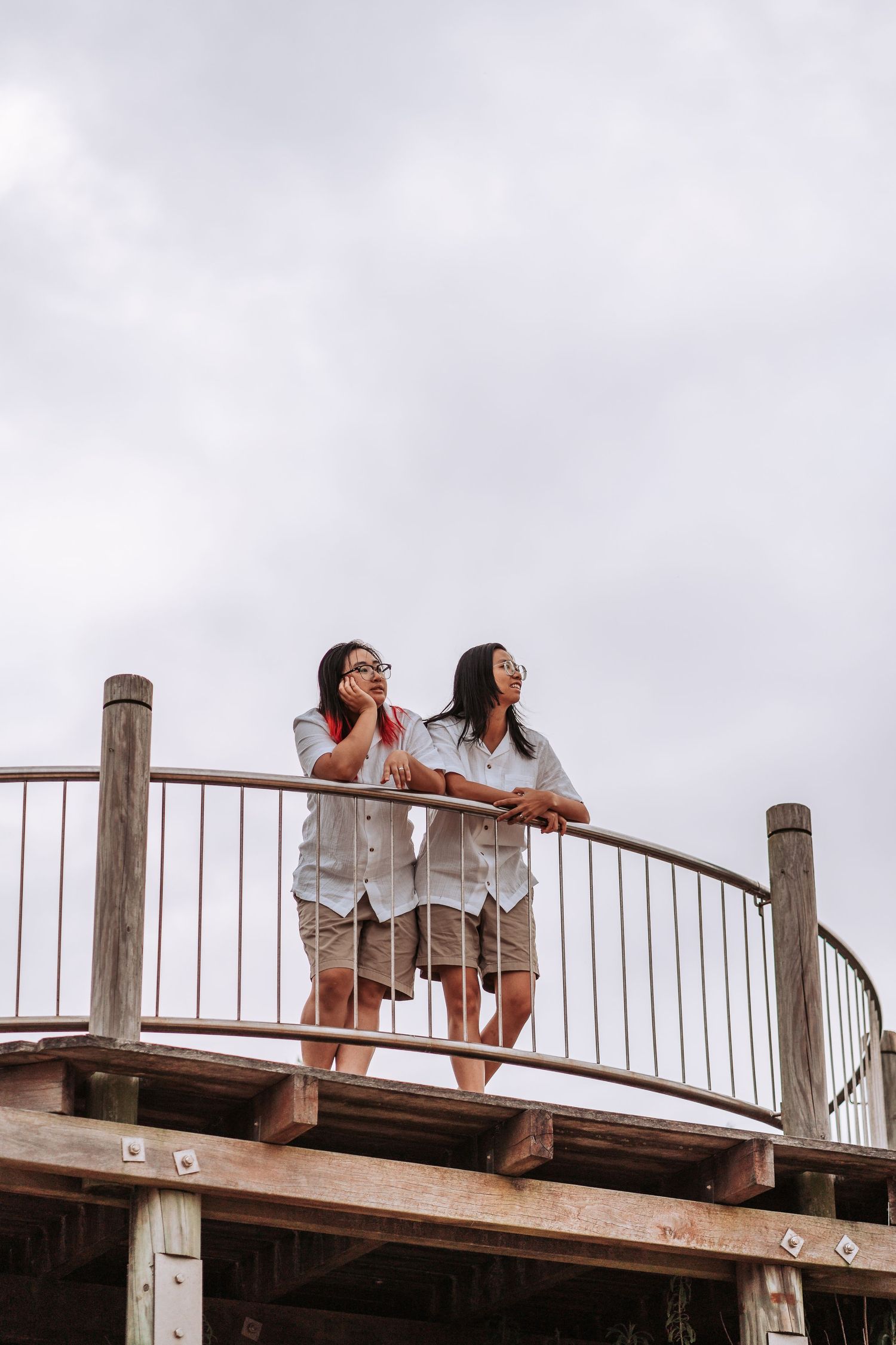 Two people wearing matching outfits share a laugh while standing on a wooden deck overlook against a cloudy sky.