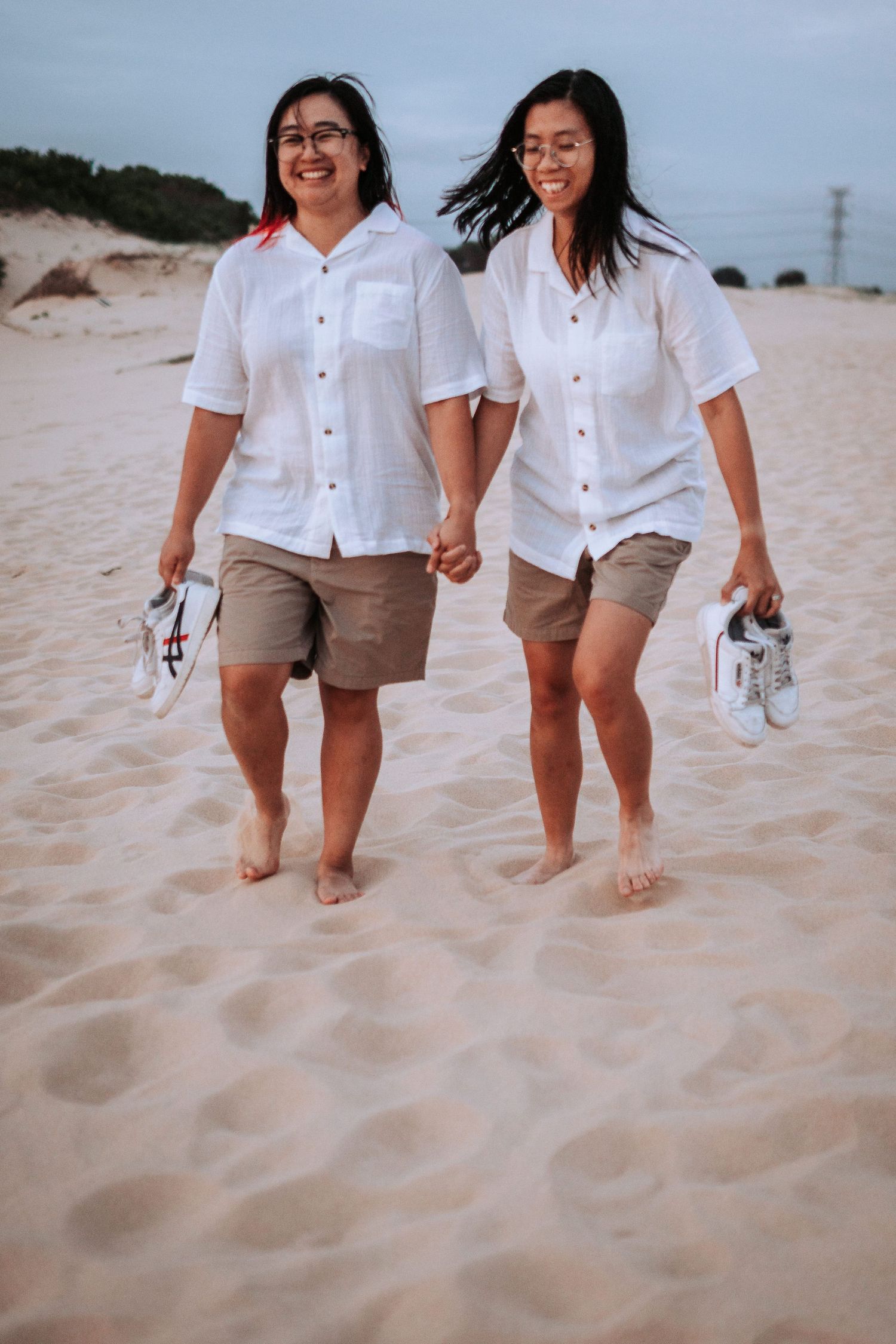 Two people holding hands walking barefoot on beach sand wearing matching white shirts and khaki shorts.