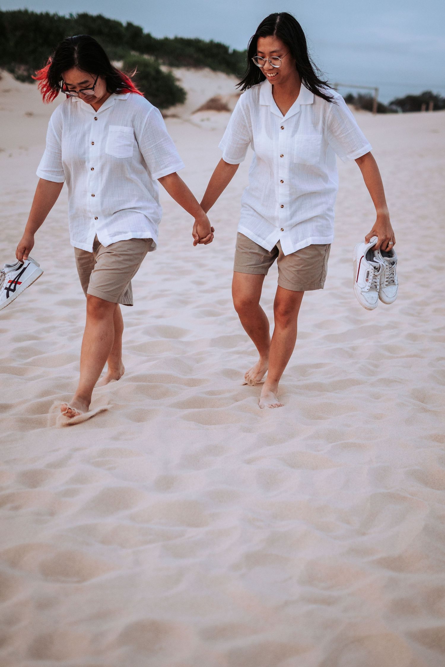 Two people in matching white shirts and khaki shorts walk barefoot on a sandy beach while holding shoes and hands.