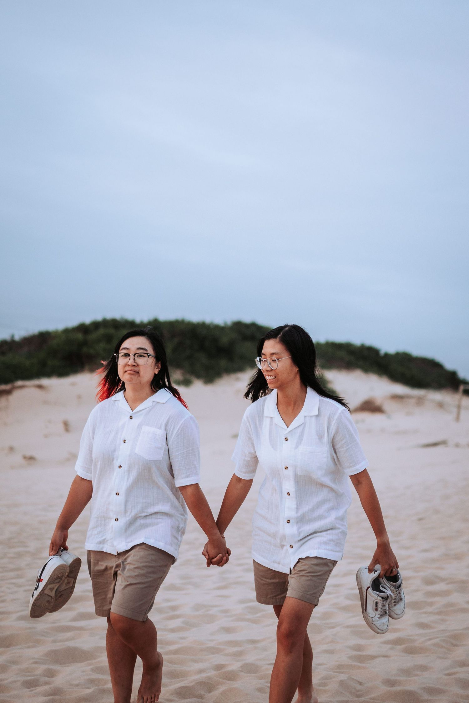 Two people in white shirts and shorts hold hands while walking barefoot along a sandy beach at dusk.